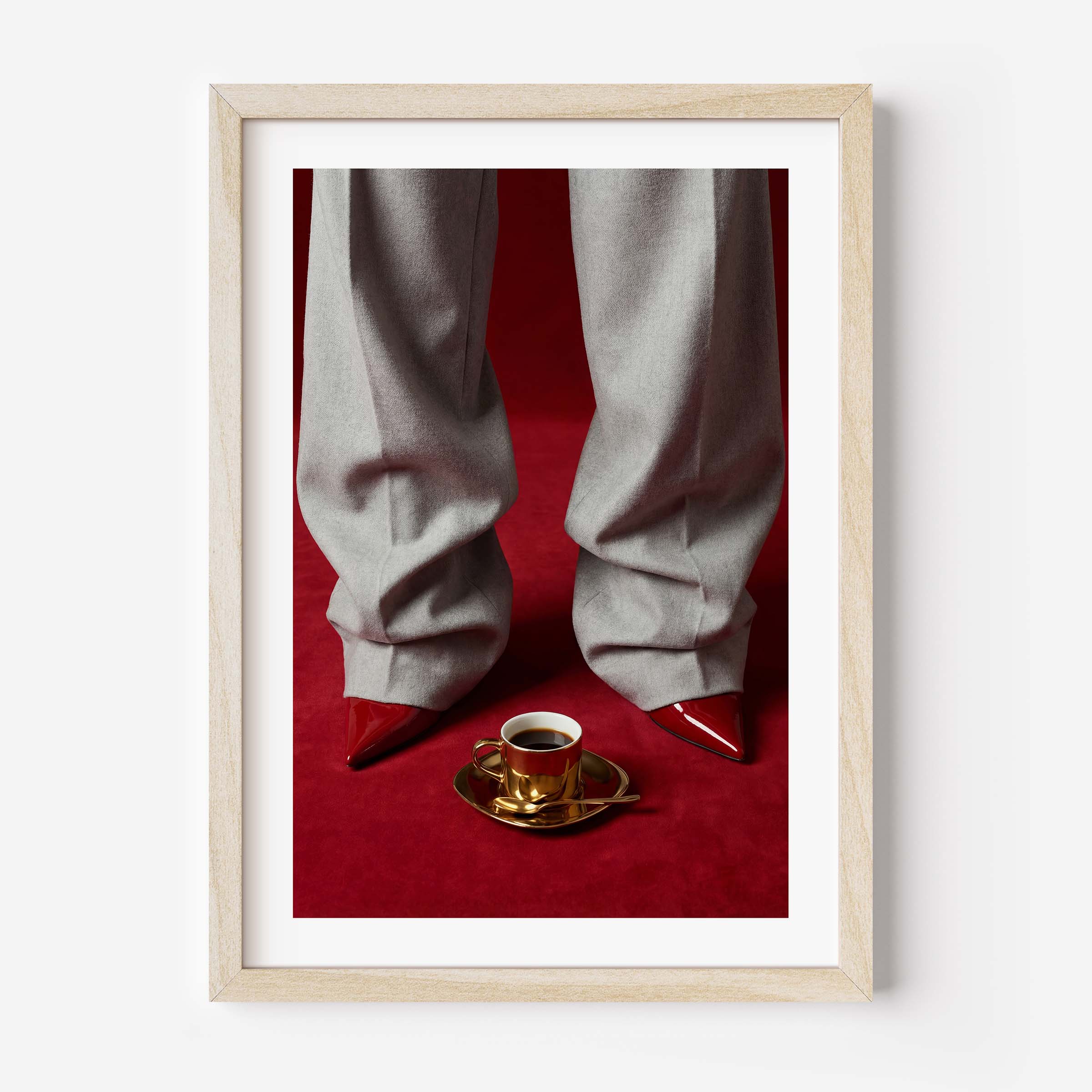 Fashion editorial wall art print showing grey wide-leg trousers, red pointed-toe stiletto heels and a gold espresso cup on a deep red velvet floor, in a black frame with white mat border.