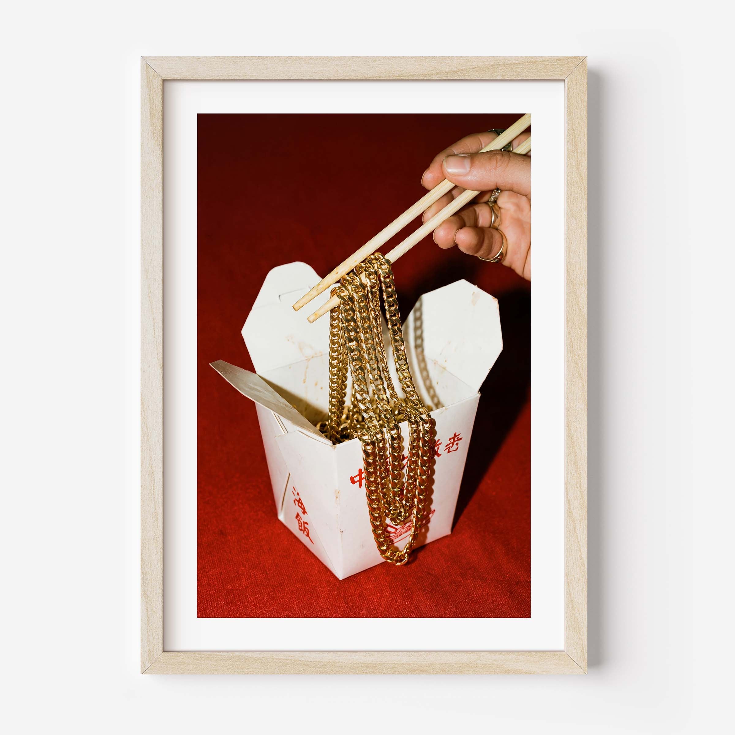 Conceptual photography print of a hand lifting gold chains with chopsticks from a Chinese takeout box on a red background, framed in black