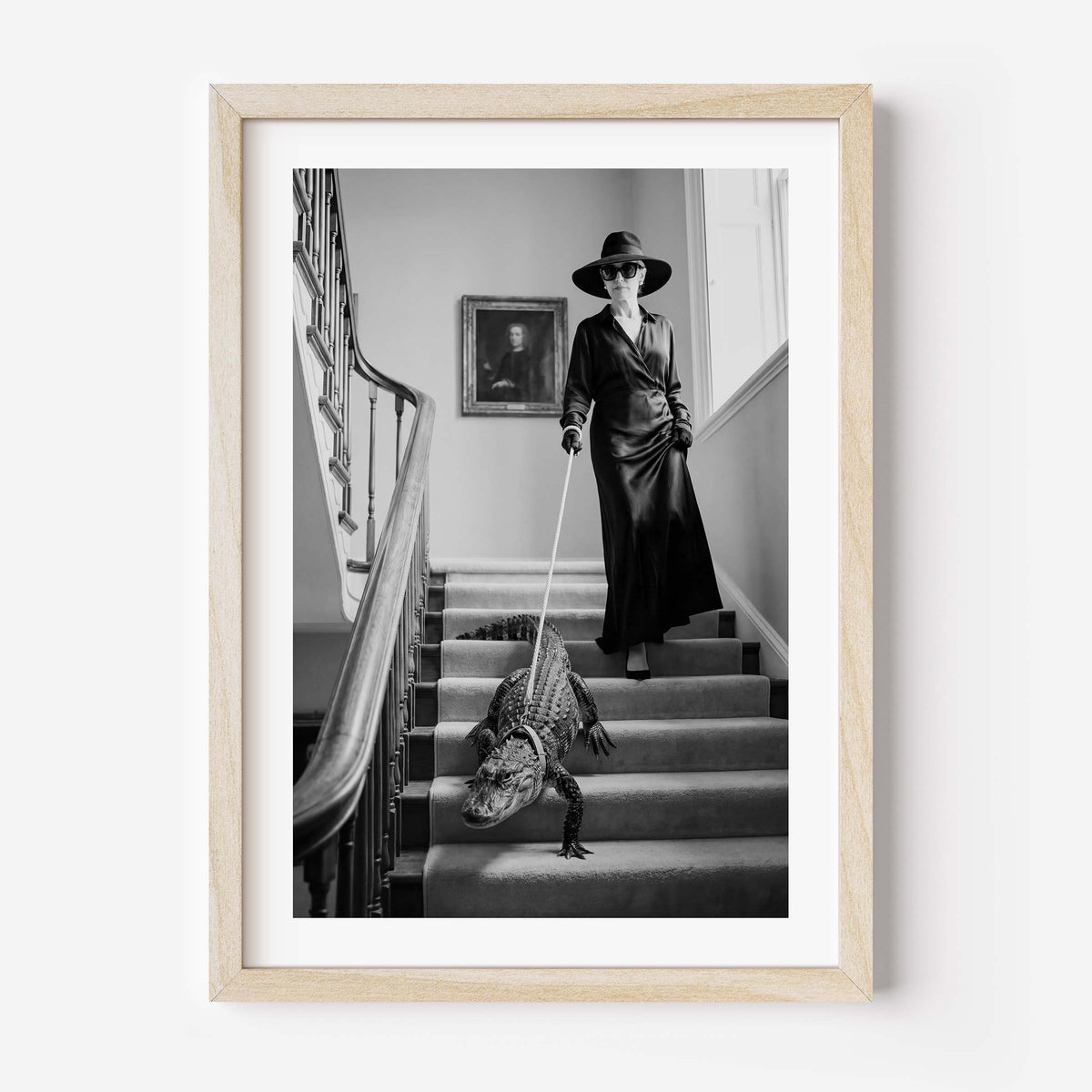A framed black and white art print depicting a woman in a long coat and hat walking a crocodile on a staircase, enclosed in a light wood frame against a white wall.