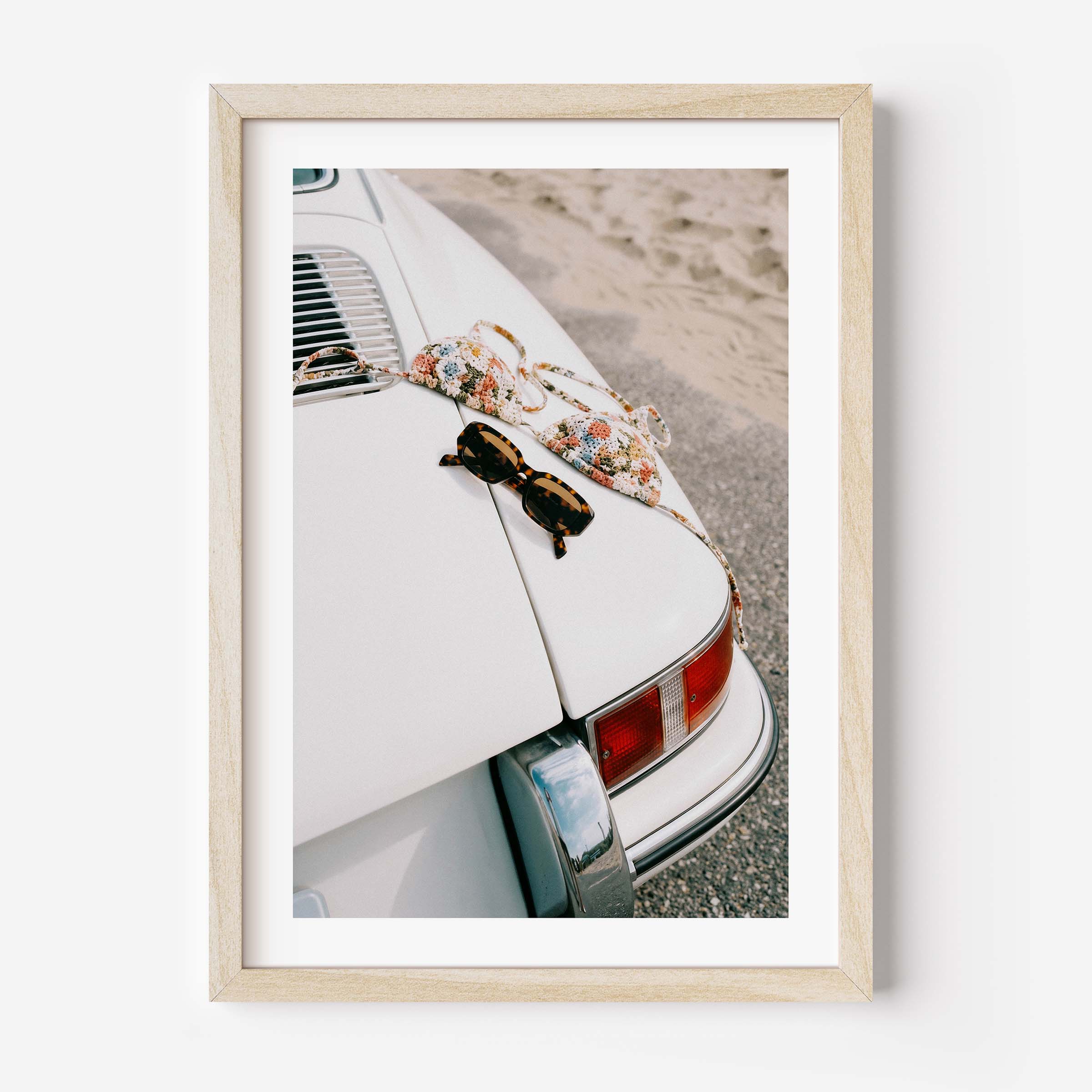 Framed wall art print showing a vintage white porsche near the beach with sunglasses and a patterned bikini placed on the back of the car.
