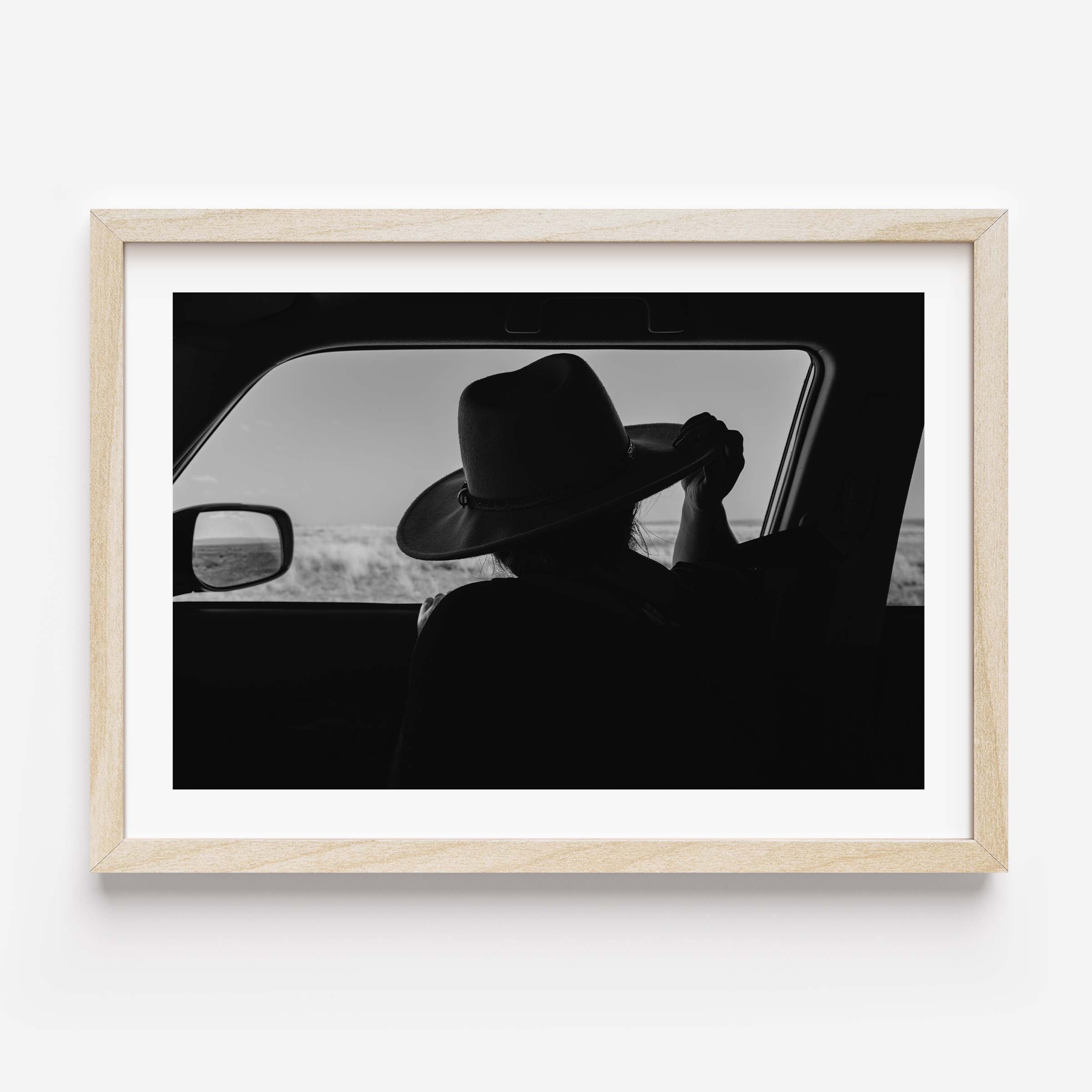 Black-and-white fine art print of a person wearing a cowboy hat seen from inside a car, looking out over an open landscape.