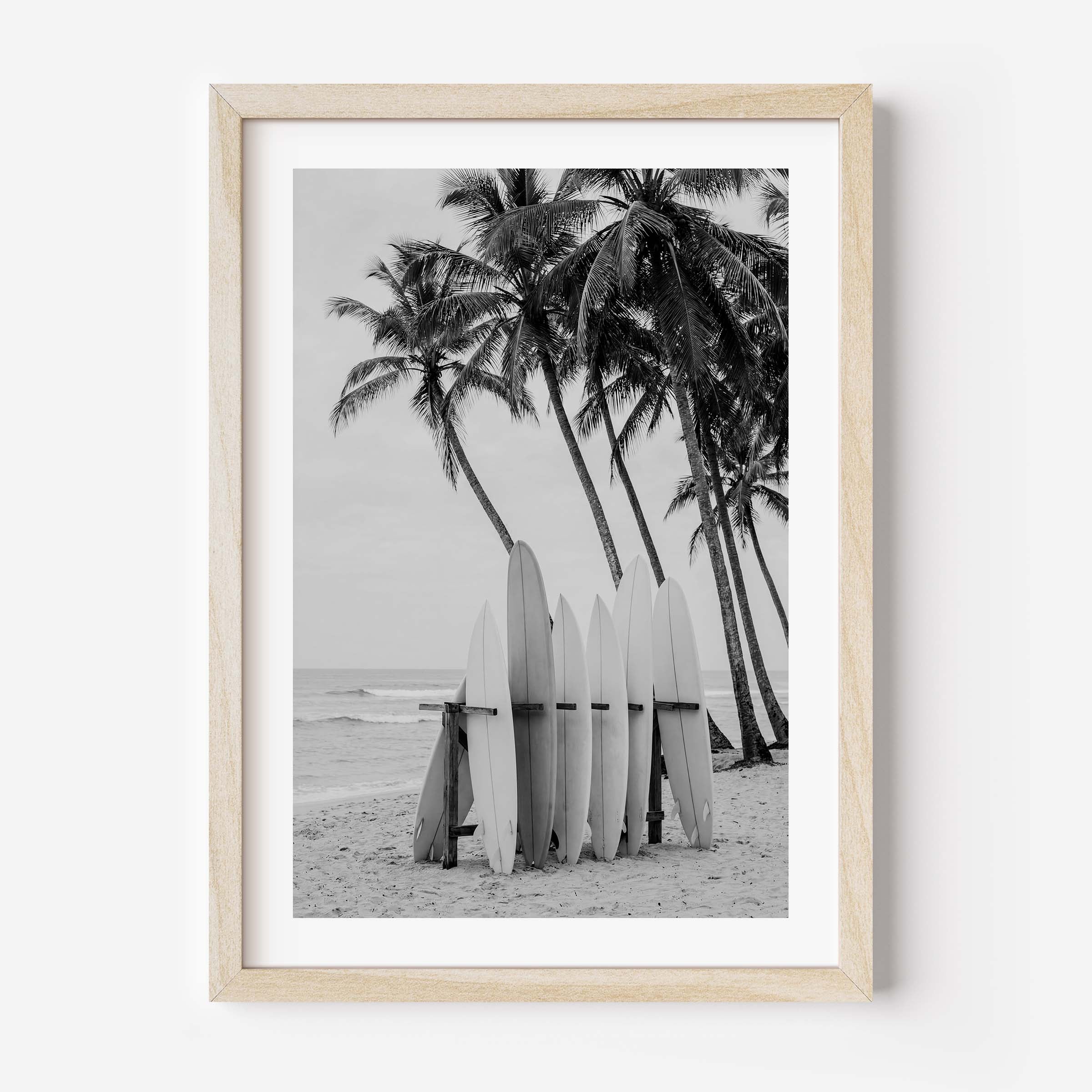Framed black and white beach photograph featuring palm trees, surfboards, and ocean waves, displayed in a light natural wood frame against a white wall.