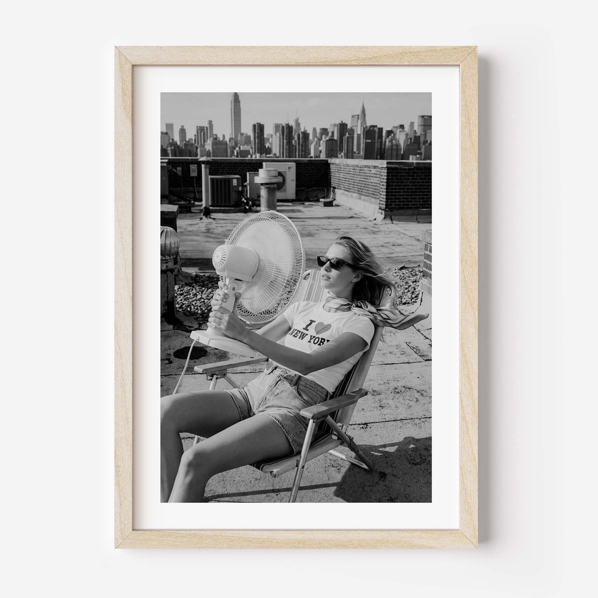 A framed black and white art print featuring a woman sitting on a rooftop chair with a fan, holding a glass of water, with a city skyline in the background. The print is housed in a light natural wood frame with a white mat, shown against a plain white wall.