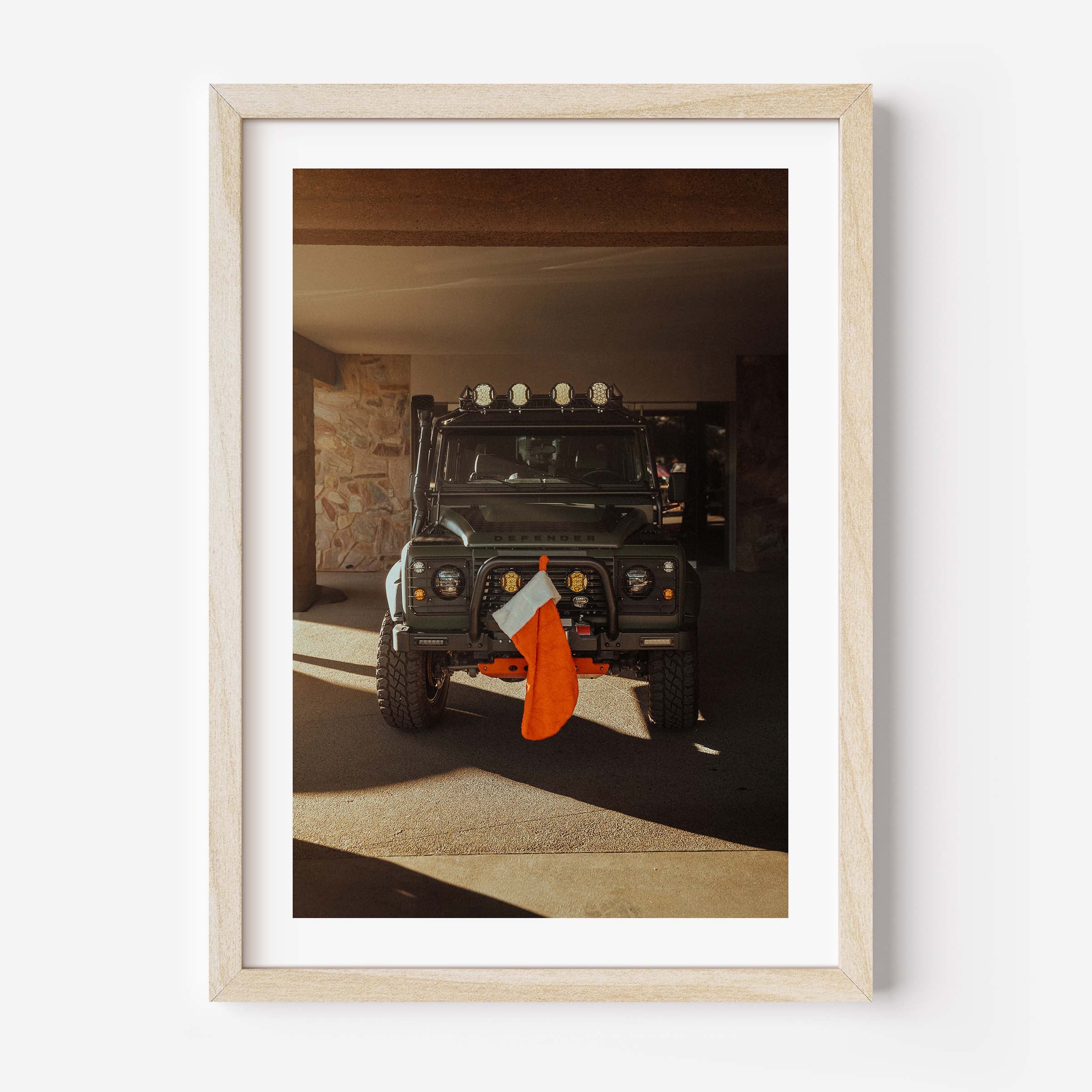 Fine art photo of a Land Rover Defender parked in a garage with warm sunlight, featuring a red Christmas stocking hanging from the front bumper.