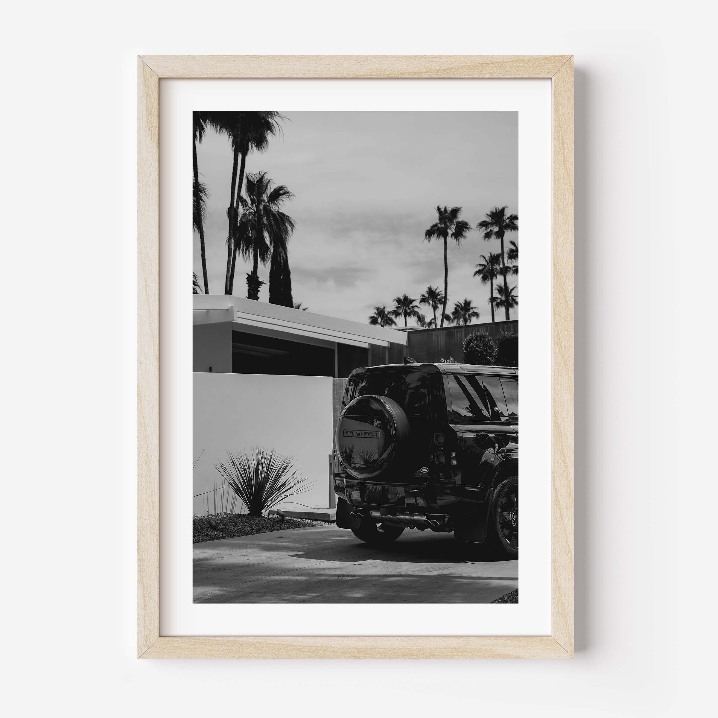 Black-and-white photo of a Land Rover Defender parked in a driveway beside a modern home, with tall palm trees and a bright sky in the background.