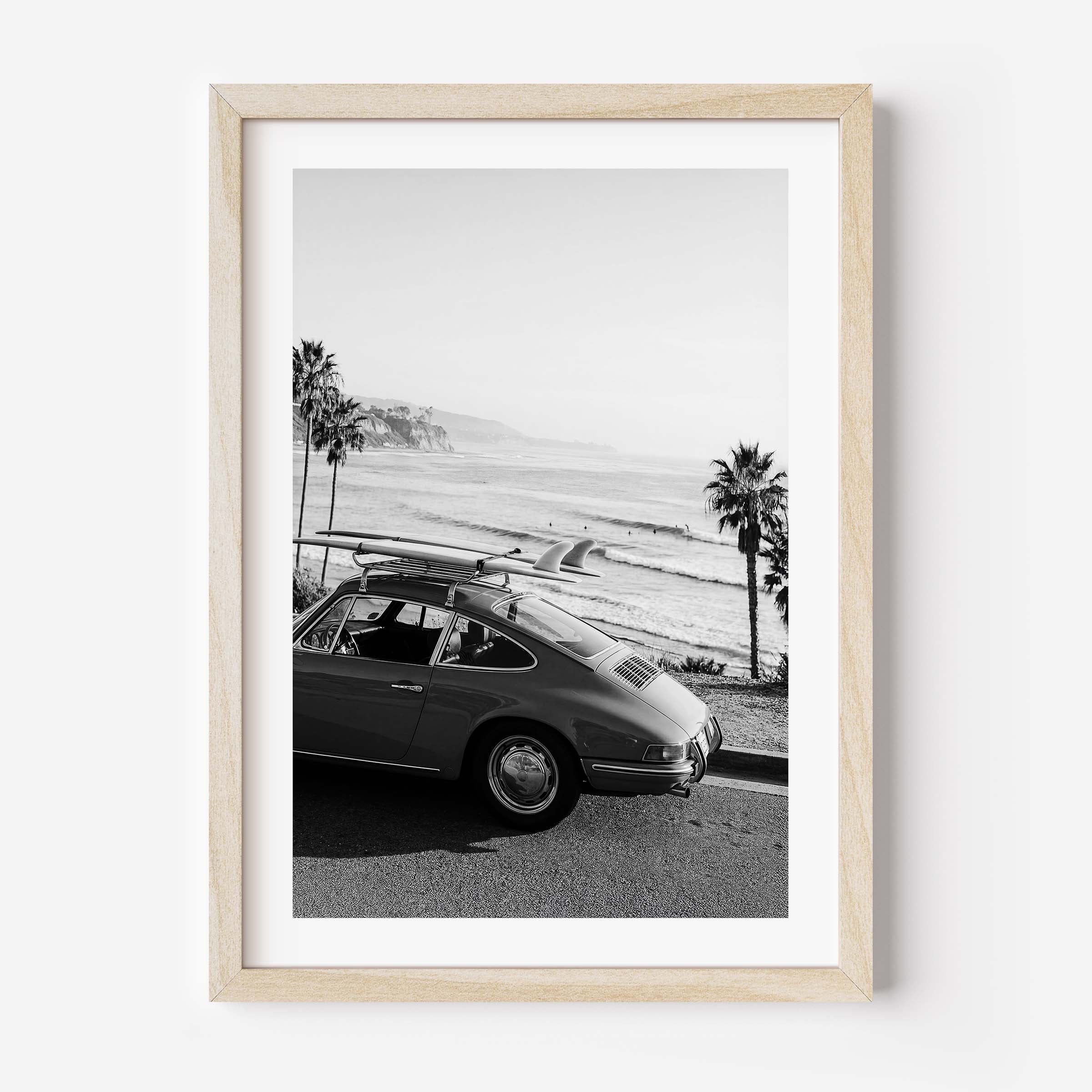Black and white framed wall art print showing a classic porsche car with surfboards parked by the ocean with palm trees and coastline in the background.