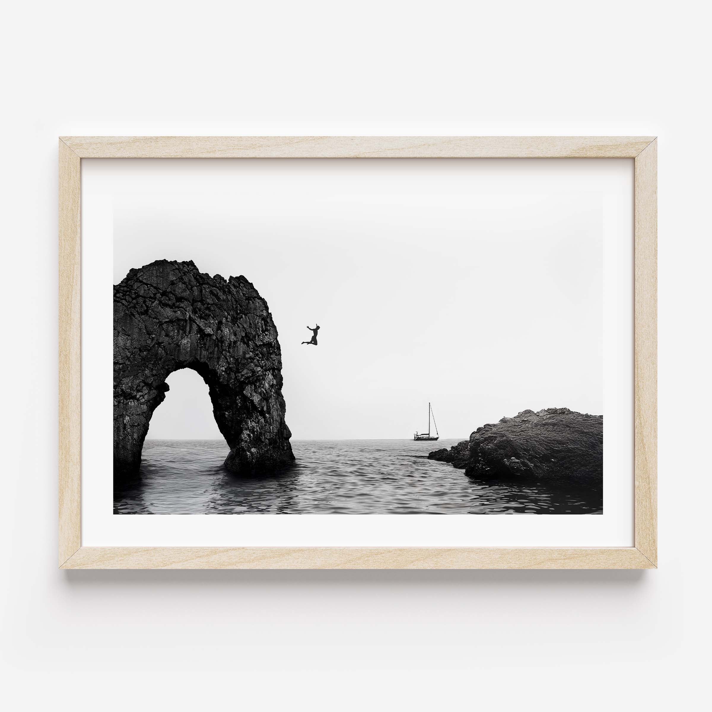 Black and white framed wall art print showing a person jumping into the ocean near a large rock arch with a sailboat in the distance.