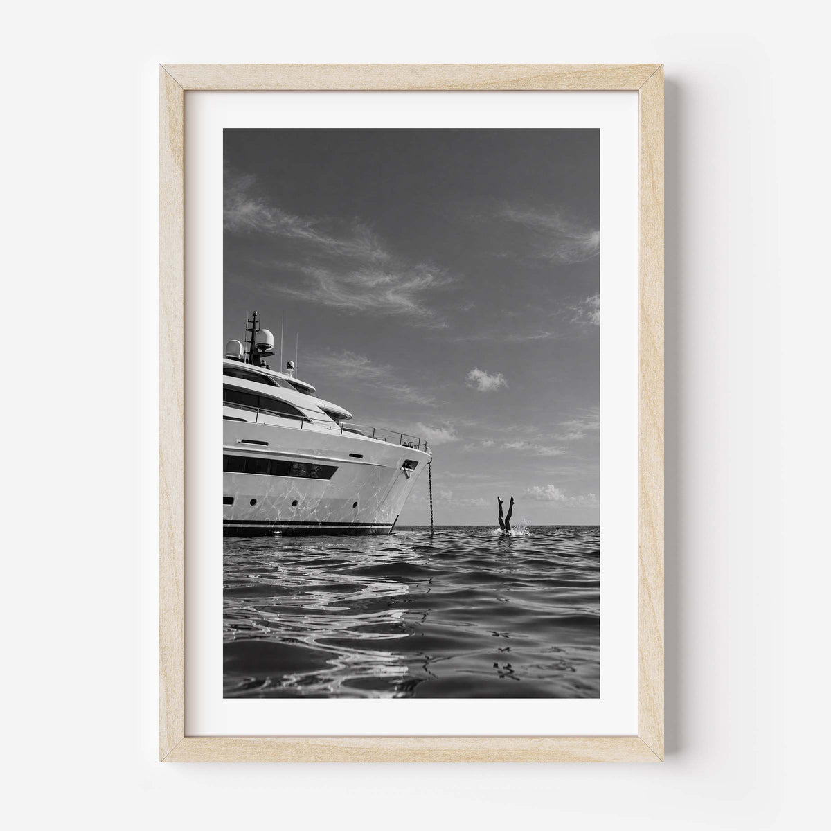 A framed black and white art print depicting a luxury yacht on calm water, with a small diver or swimmer in the distance, presented in a light natural wood frame against a white wall.