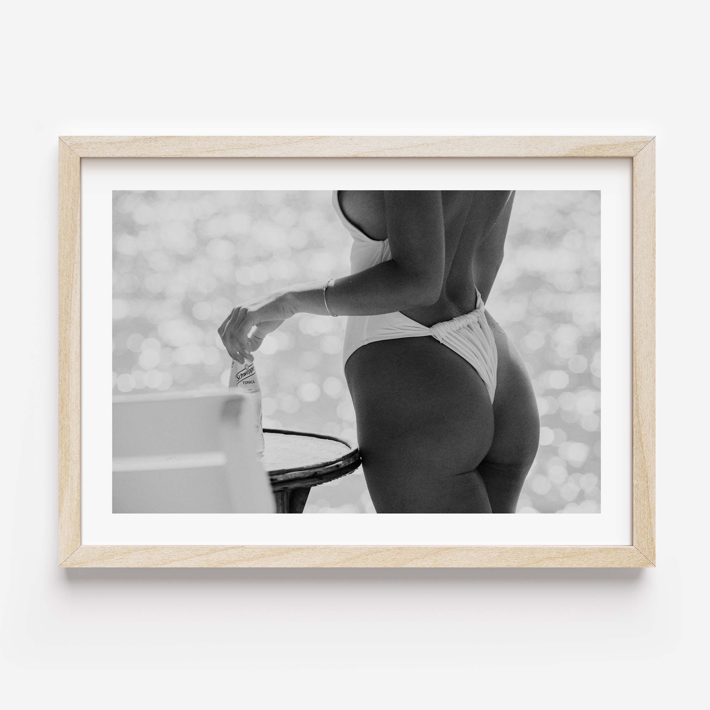 Black-and-white photo of a woman in a white one-piece holding a glass bottle by a sunlit seaside table