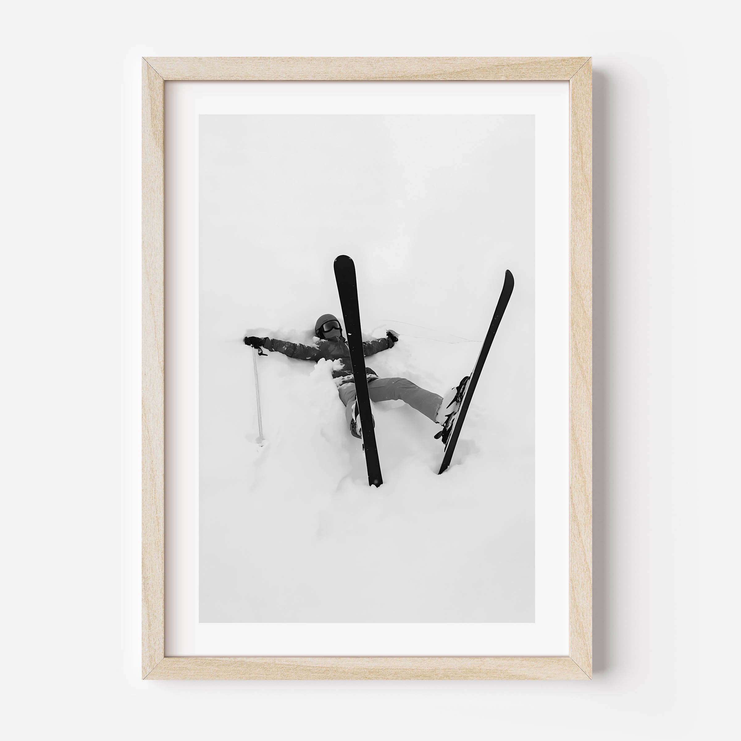 Black and white fine art print of a skier lying in deep snow with skis and pole scattered, framed in black.