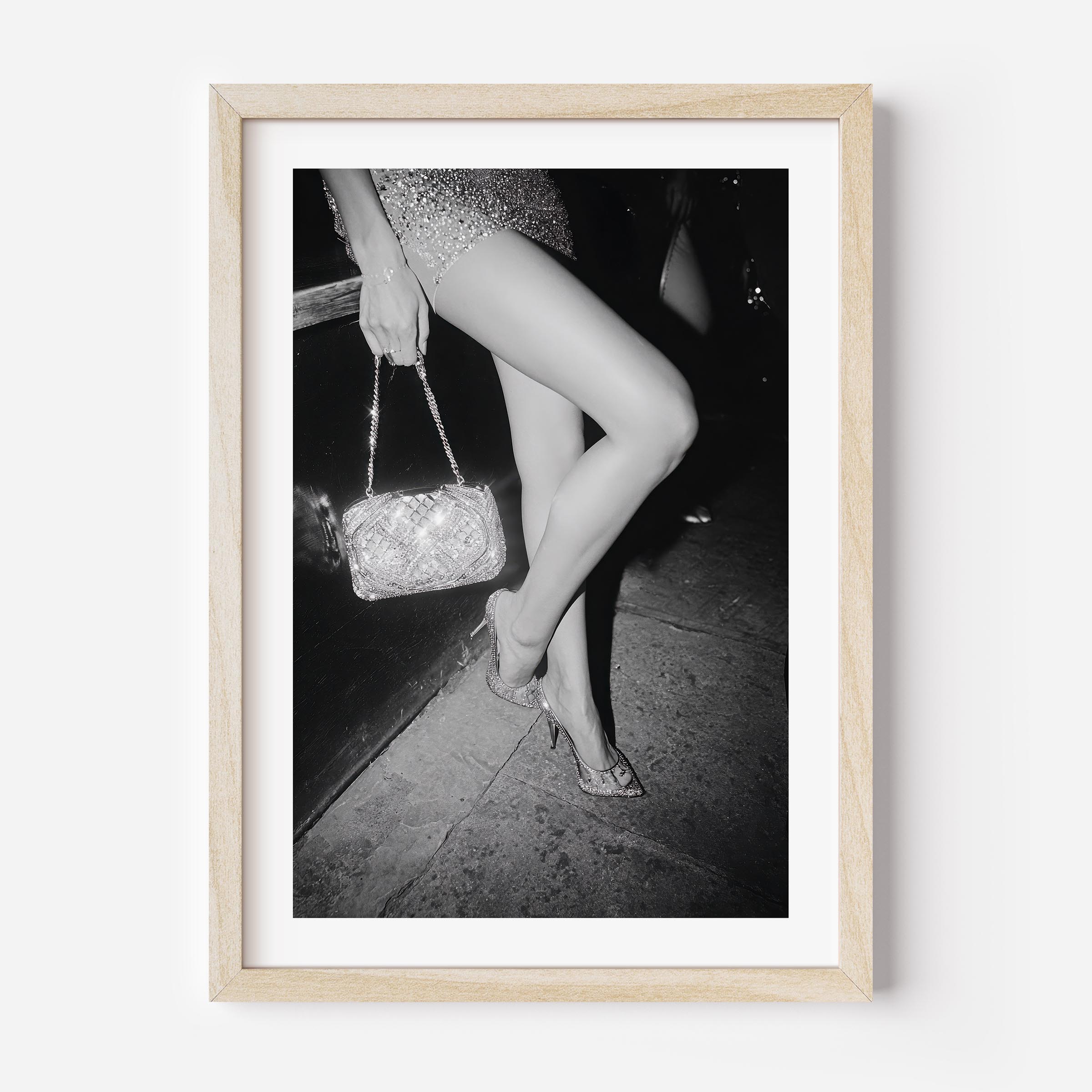 Black-and-white fine art photograph of a woman’s legs in high heels and sequin dress, holding a glittering handbag on a city sidewalk.