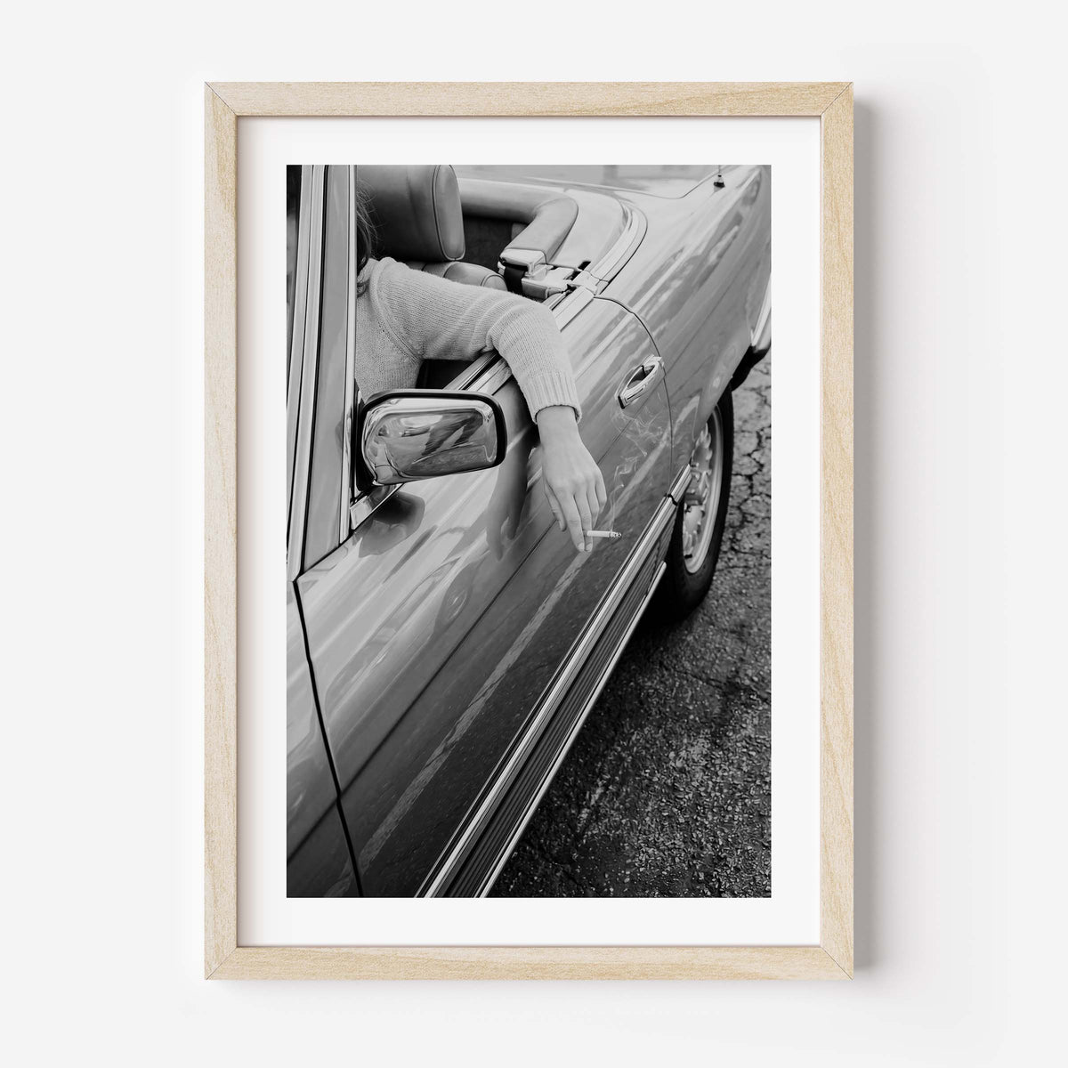 A framed black and white art print depicting a person in a convertible car with their arm out the window, held by a light natural wood frame and white matting, shown against a plain white wall.