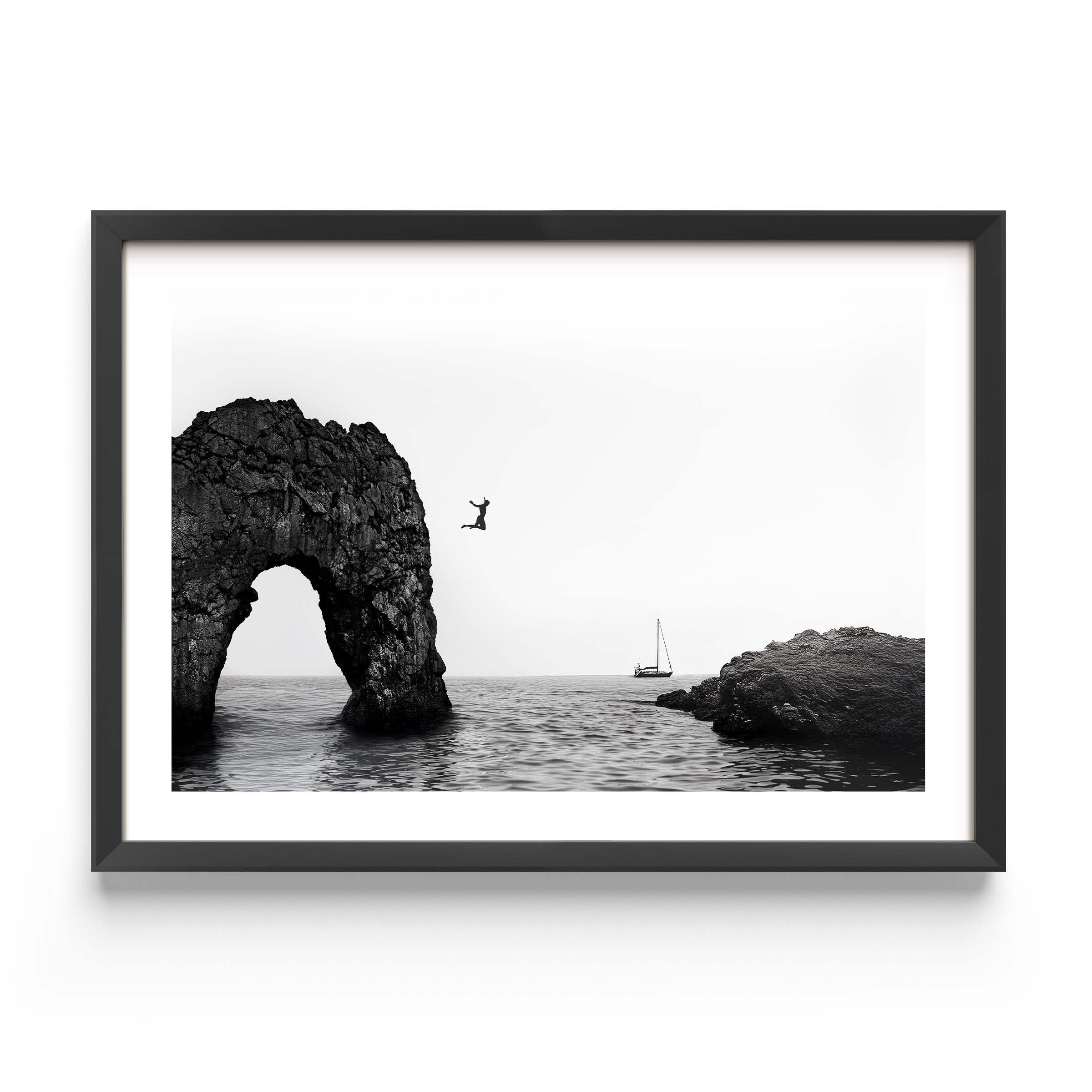 Black and white framed wall art print showing a person jumping into the ocean near a large rock arch with a sailboat in the distance.