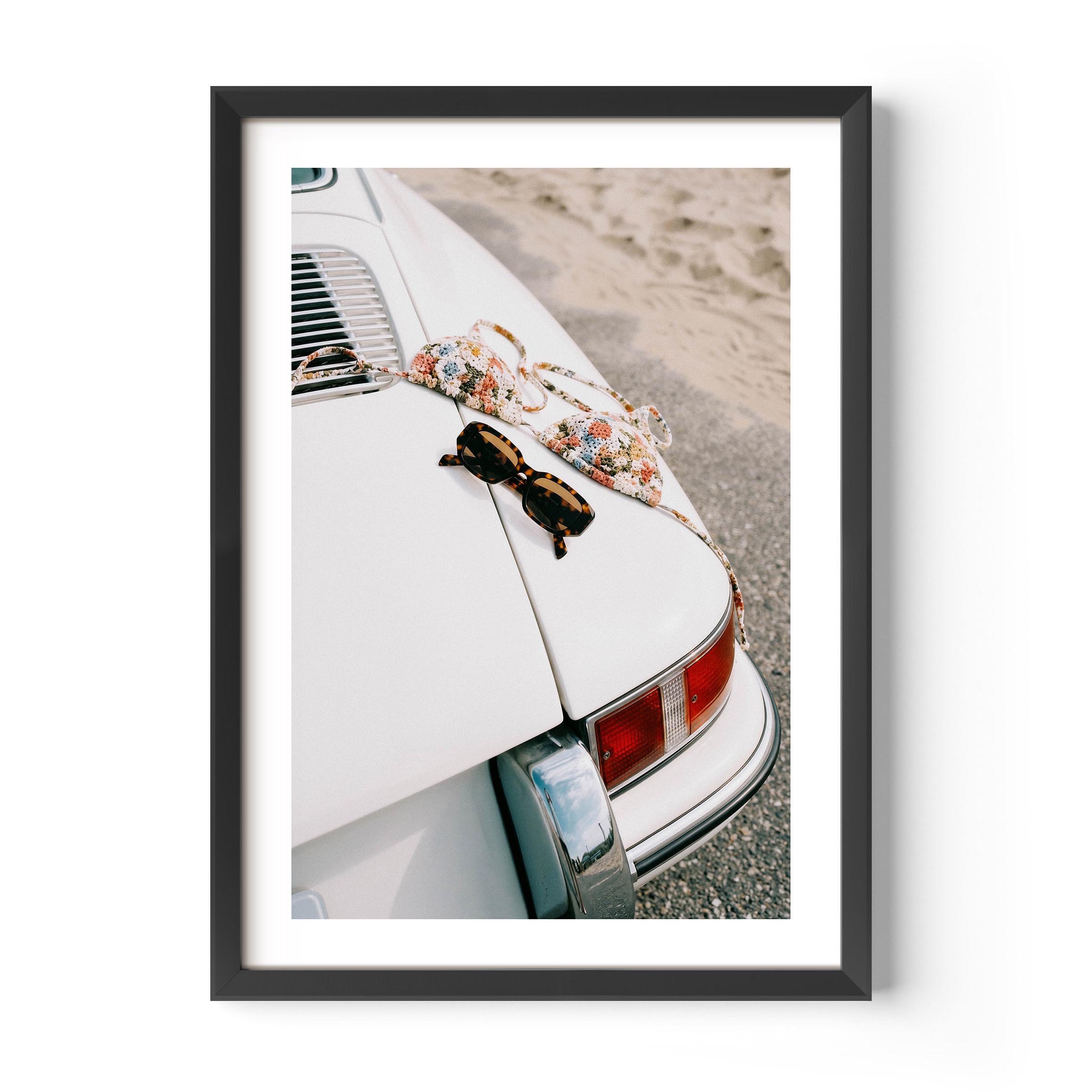 Framed wall art print showing a vintage white porsche near the beach with sunglasses and a patterned bikini placed on the back of the car.