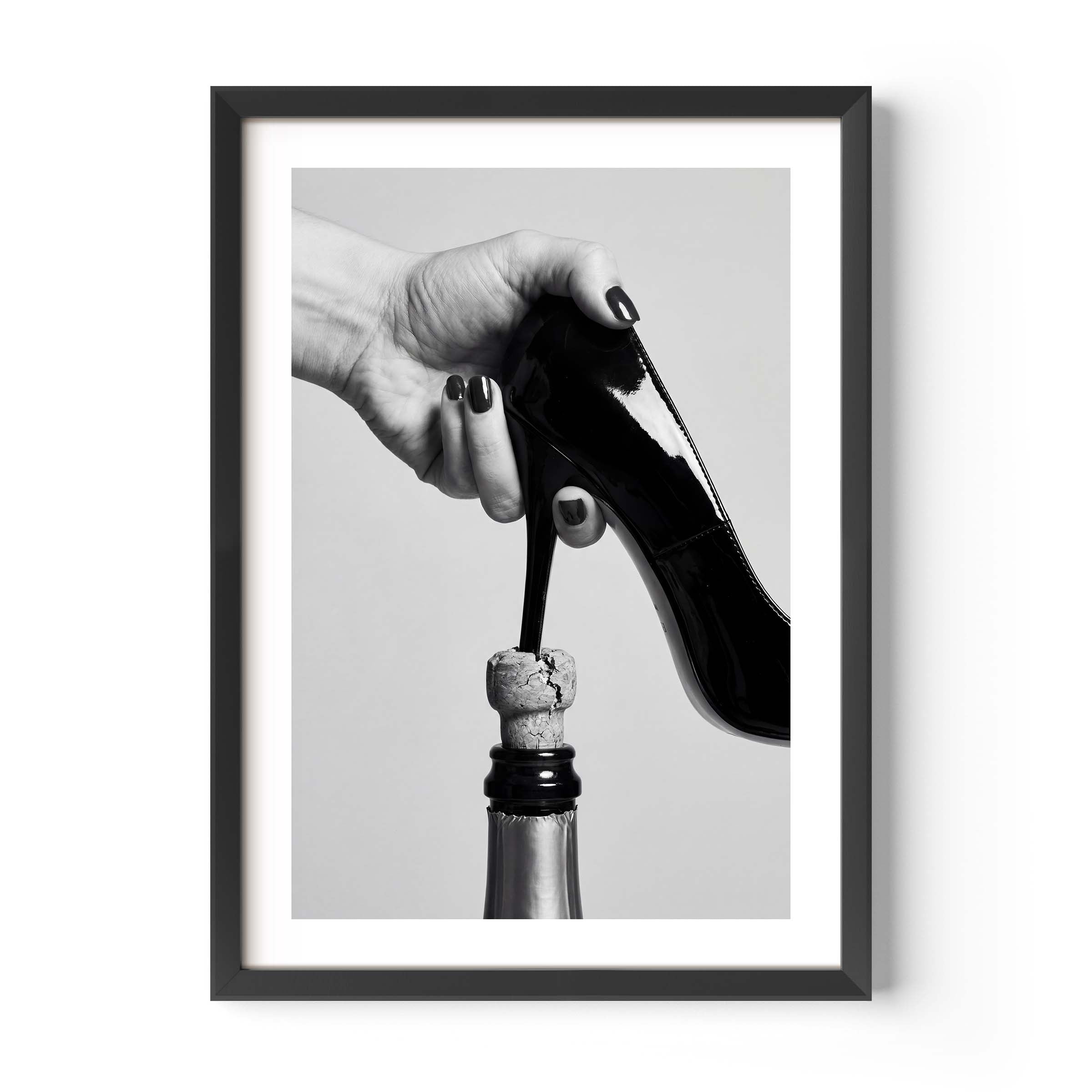 A framed black and white art print featuring a hand holding a high-heeled shoe above a champagne bottle, displayed on a clean white background.