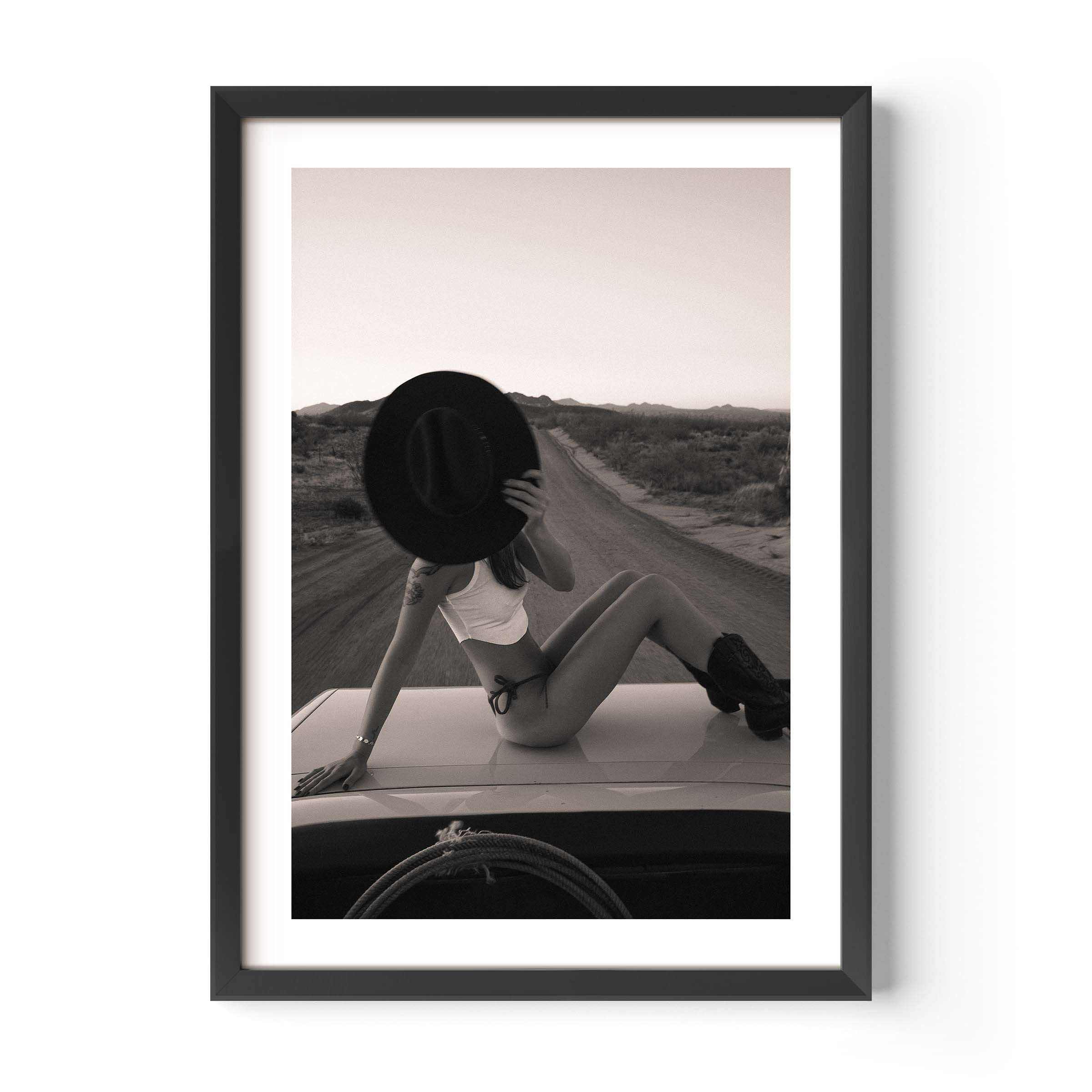 Fine art photo print of a woman sitting on a car roof on a desert road, holding a wide-brim hat in front of her face at sunset.