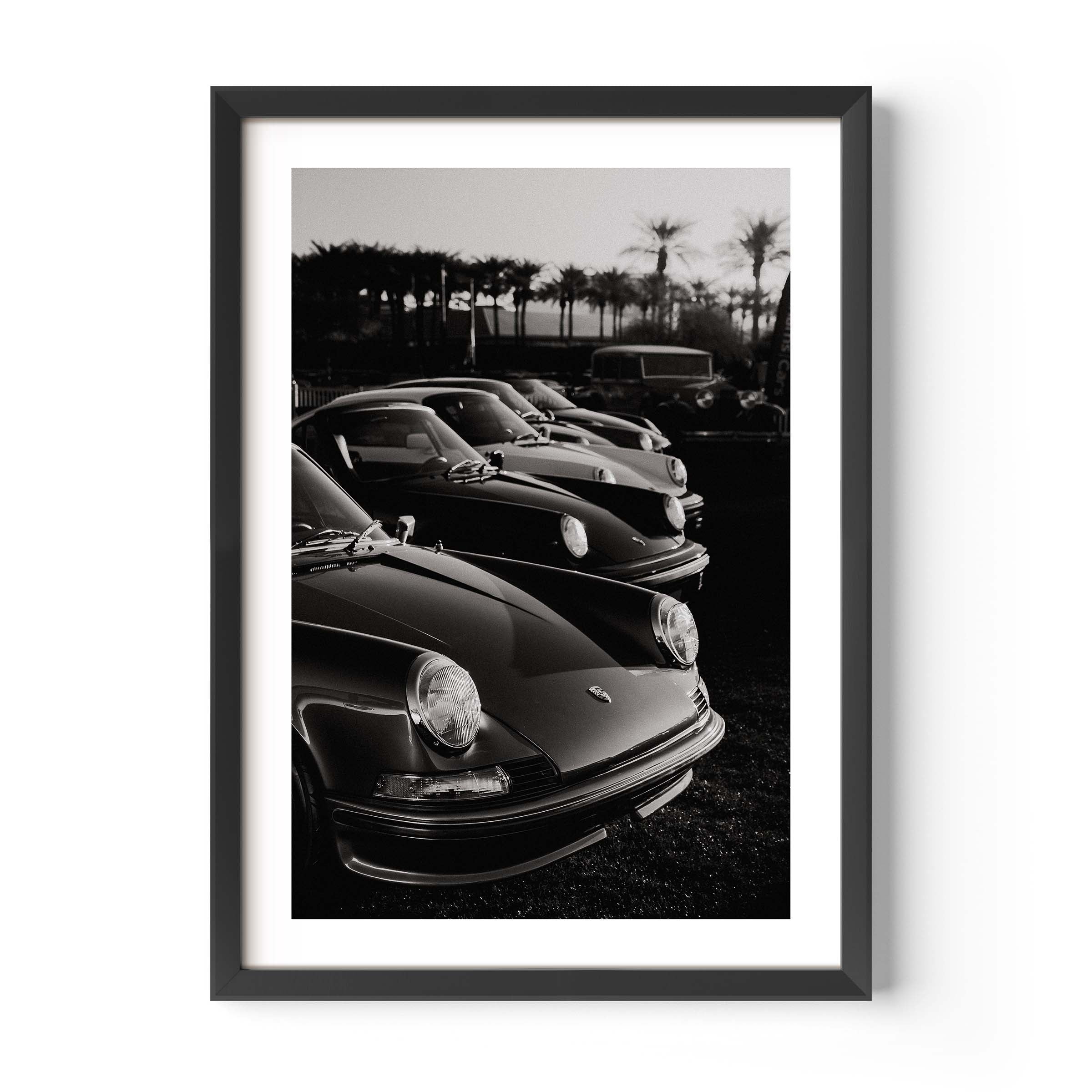 Black-and-white photo print of a row of classic Porsche 911 sports cars parked with palm trees in the background.