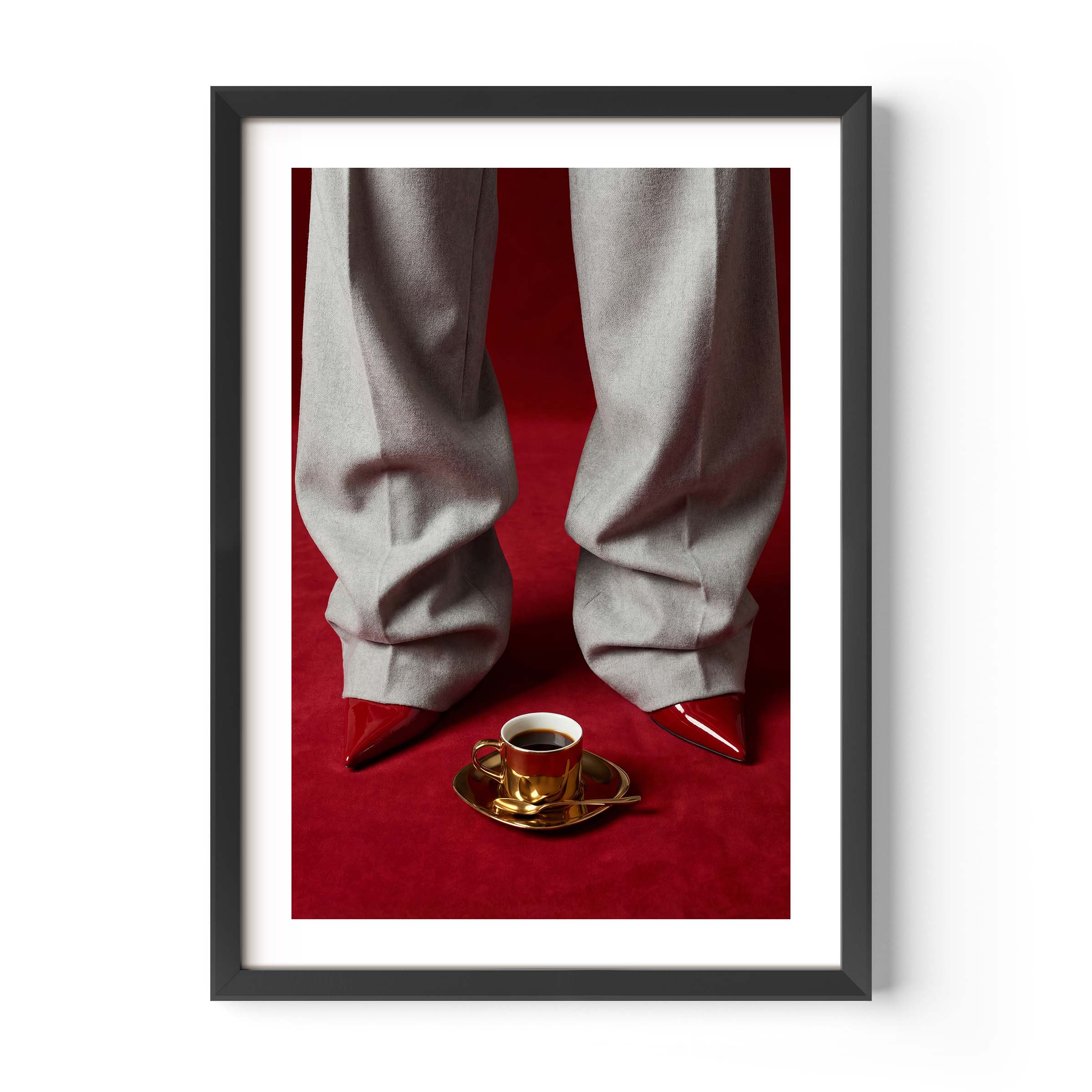 Fashion editorial wall art print showing grey wide-leg trousers, red pointed-toe stiletto heels and a gold espresso cup on a deep red velvet floor, in a black frame with white mat border.