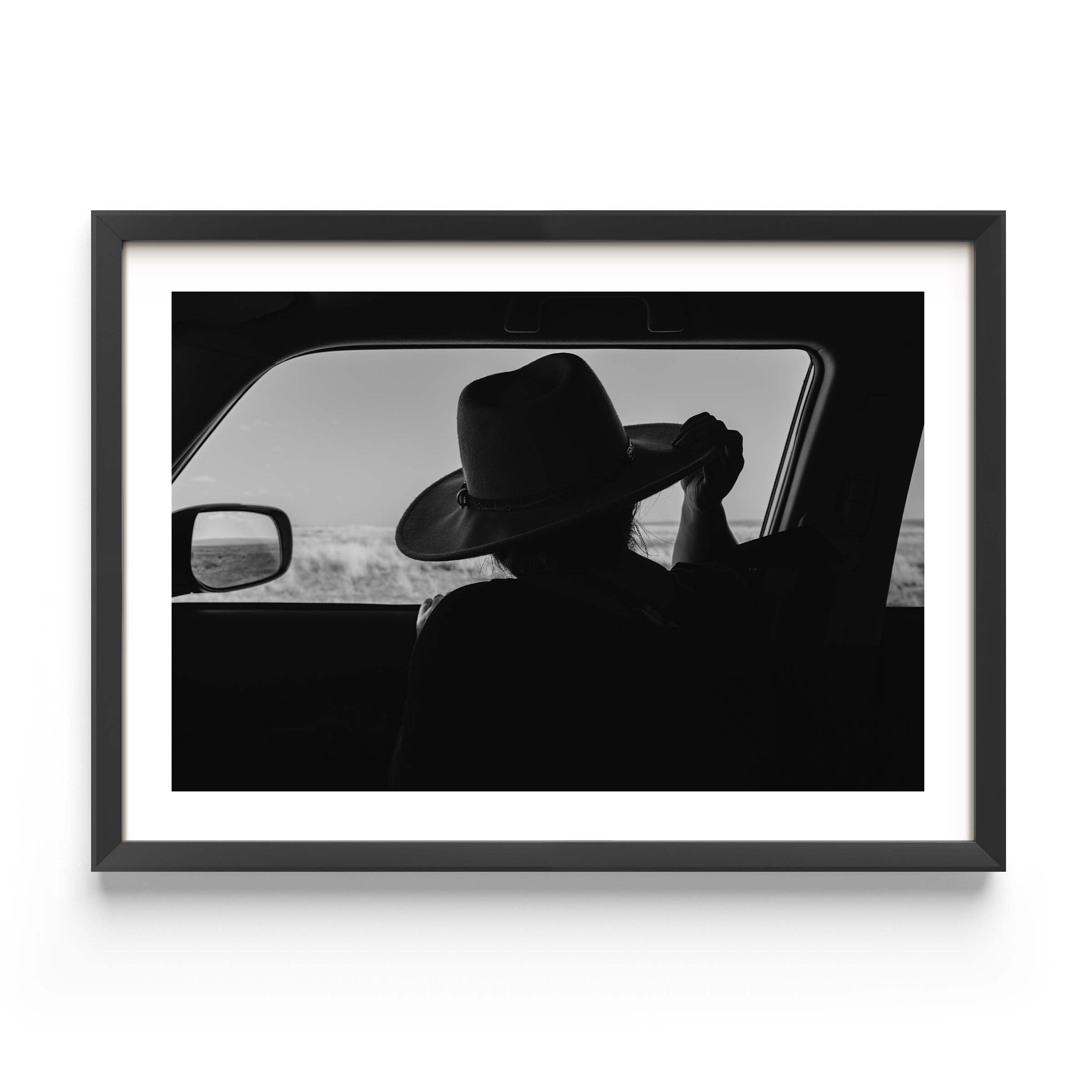 Black-and-white fine art print of a person wearing a cowboy hat seen from inside a car, looking out over an open landscape.