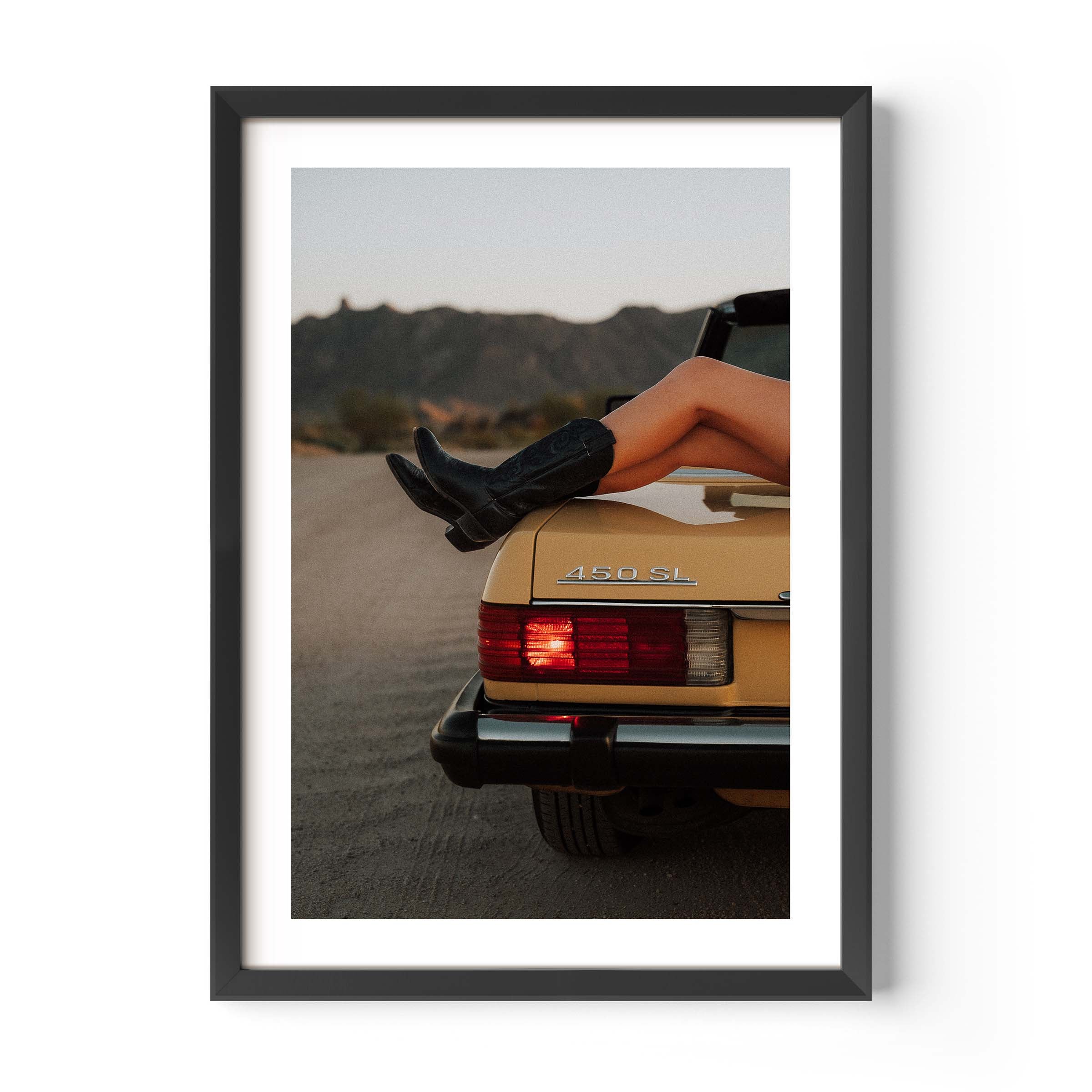 Fine art photo of the rear of a classic Mercedes 450 SL at dusk, with a person’s legs in black boots resting on the trunk and mountains in the background.