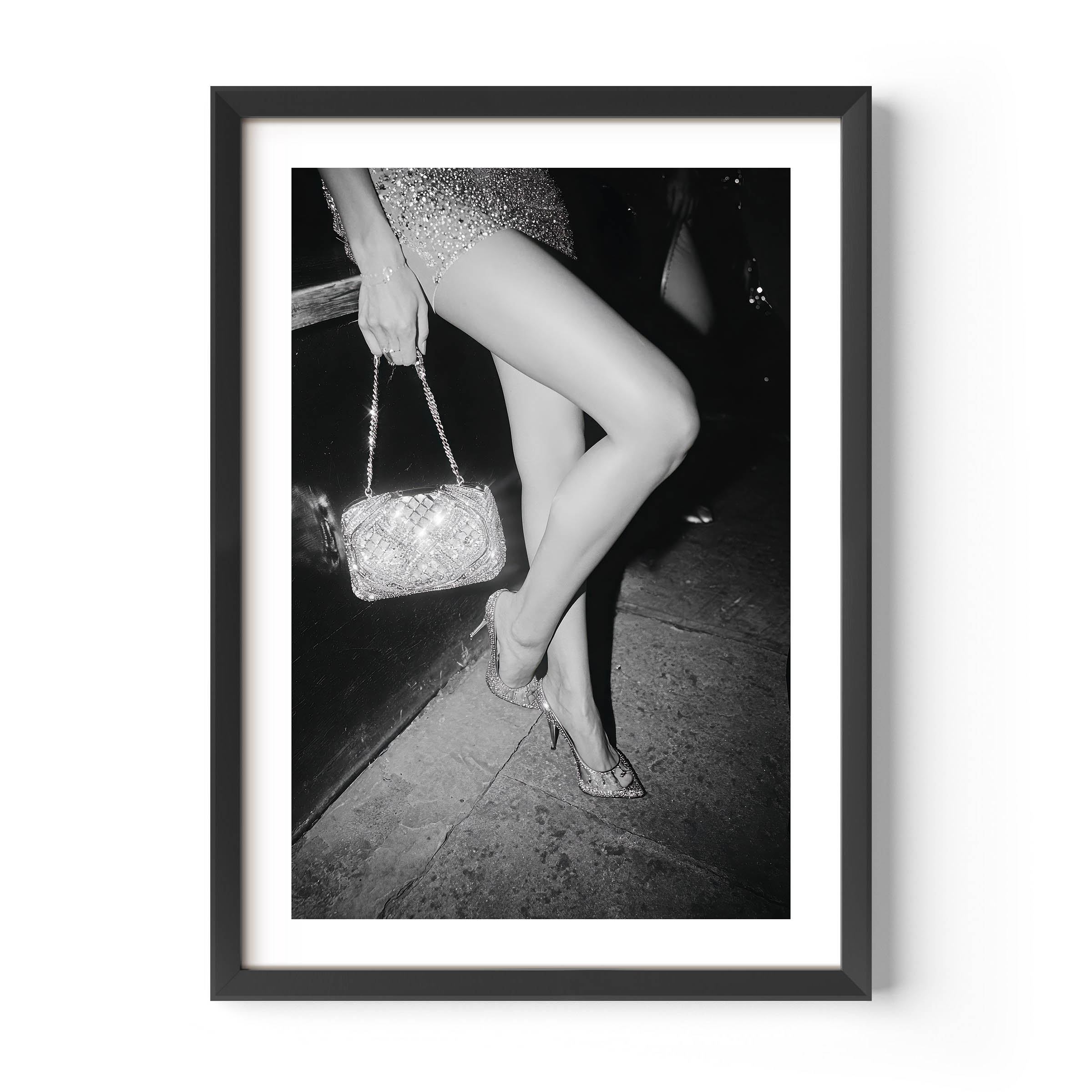 Black-and-white fine art photograph of a woman’s legs in high heels and sequin dress, holding a glittering handbag on a city sidewalk.