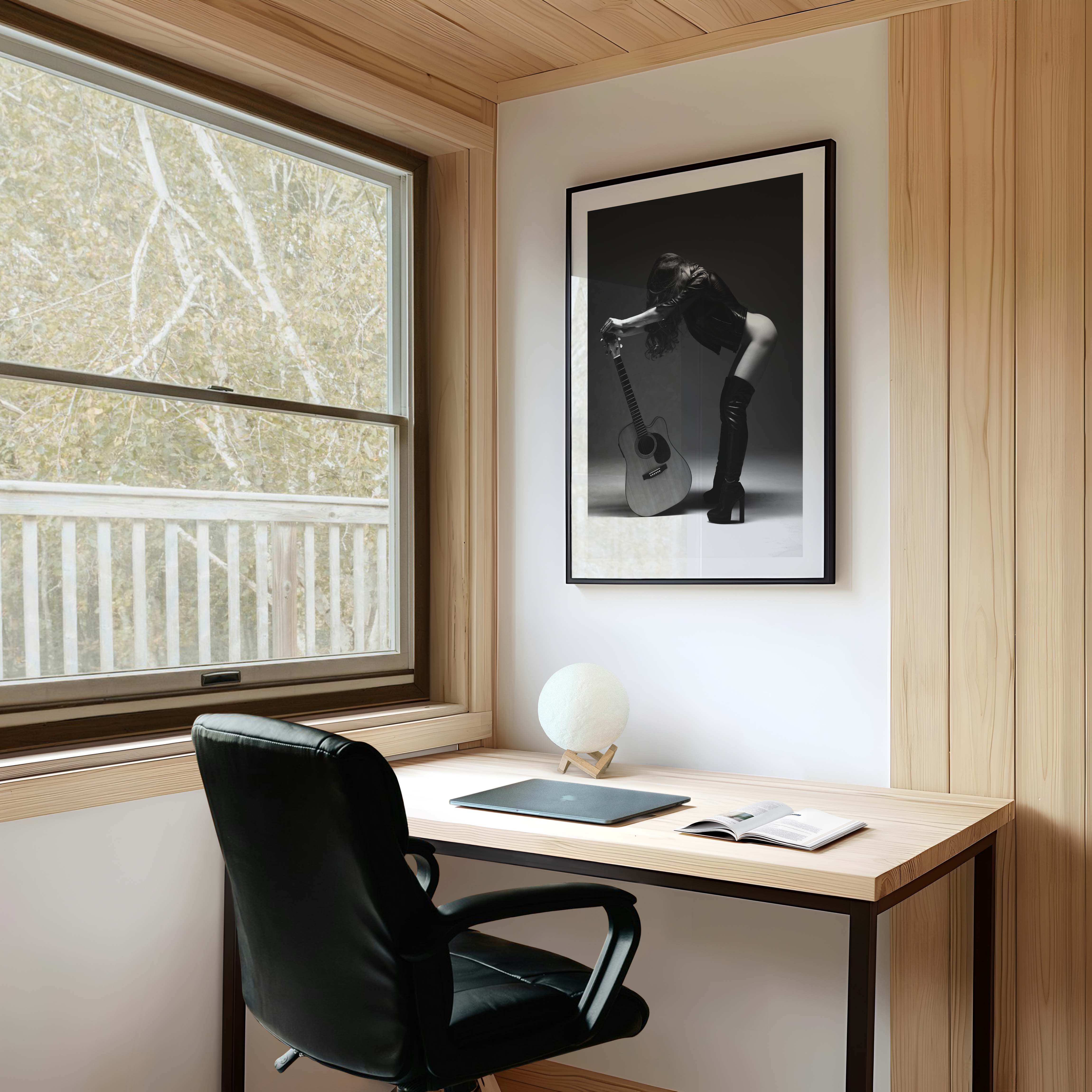 Black-and-white photo of a woman in a leather jacket and thigh-high boots leaning over an acoustic guitar in a studio, framed with white mat.