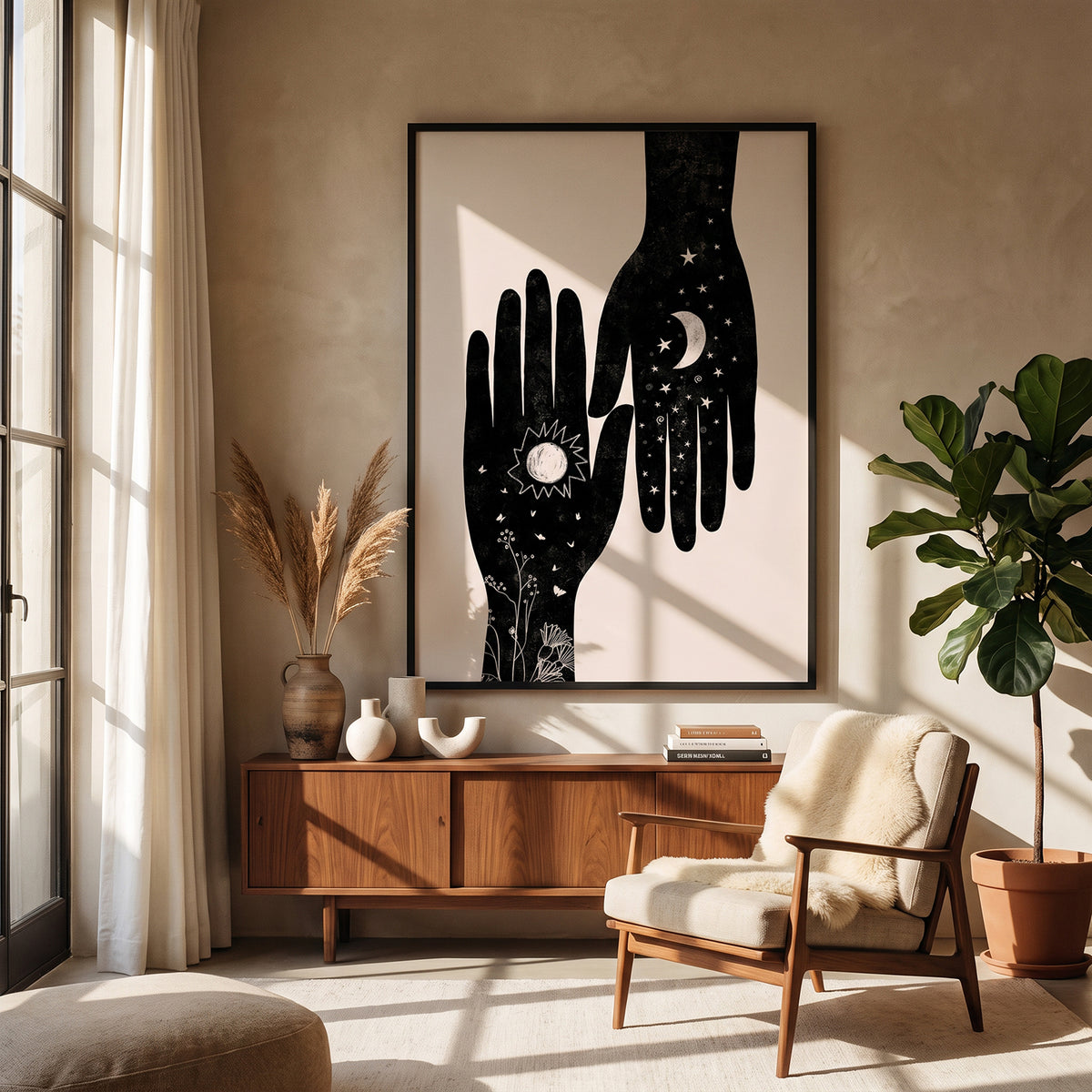 A large framed wall art print hangs above a wooden sideboard in a sunlit living room. The artwork shows two black hands with celestial and floral details on a warm off-white background.