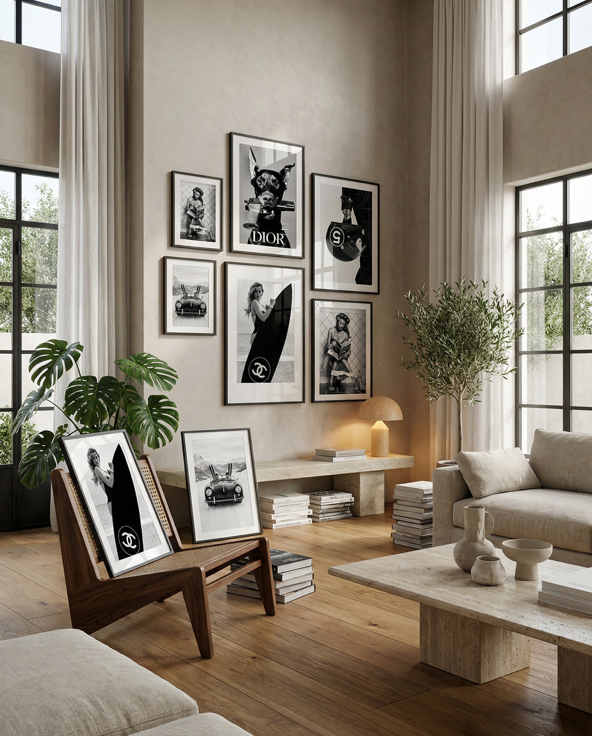 A styled interior room with a gallery wall of black-and-white framed art, a beige sofa, wood floors, plants, and a coffee table. Several framed prints are leaning on a wooden chair in the foreground.