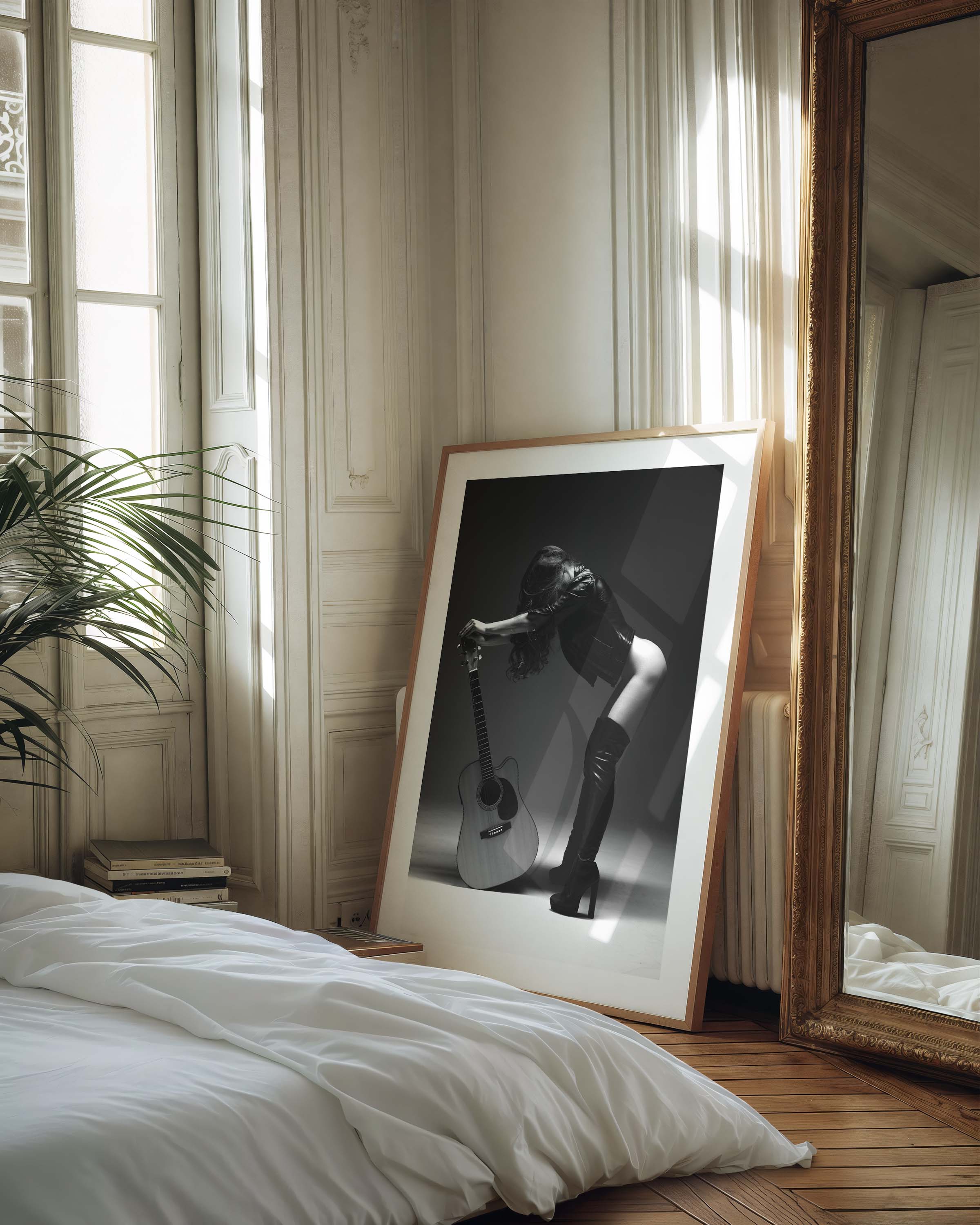 Black-and-white photo of a woman in a leather jacket and thigh-high boots leaning over an acoustic guitar in a studio, framed with white mat.