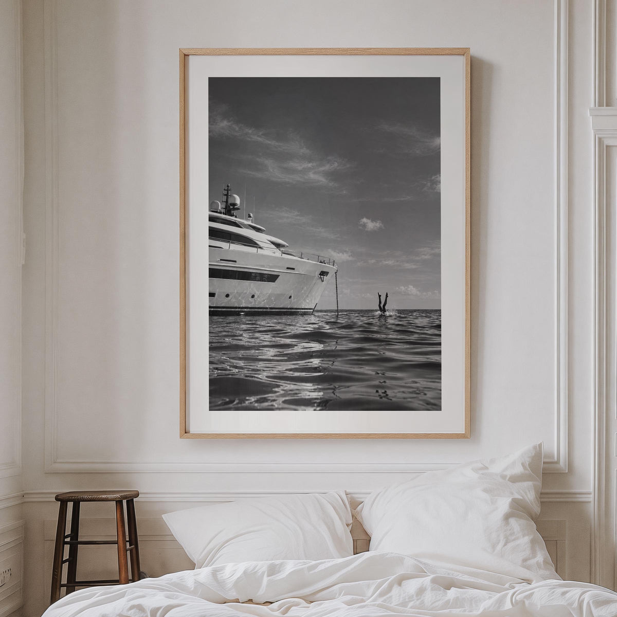 A framed black and white art print of a luxury yacht on the water, styled on a white wall above a bed with white linens and a small wooden stool.