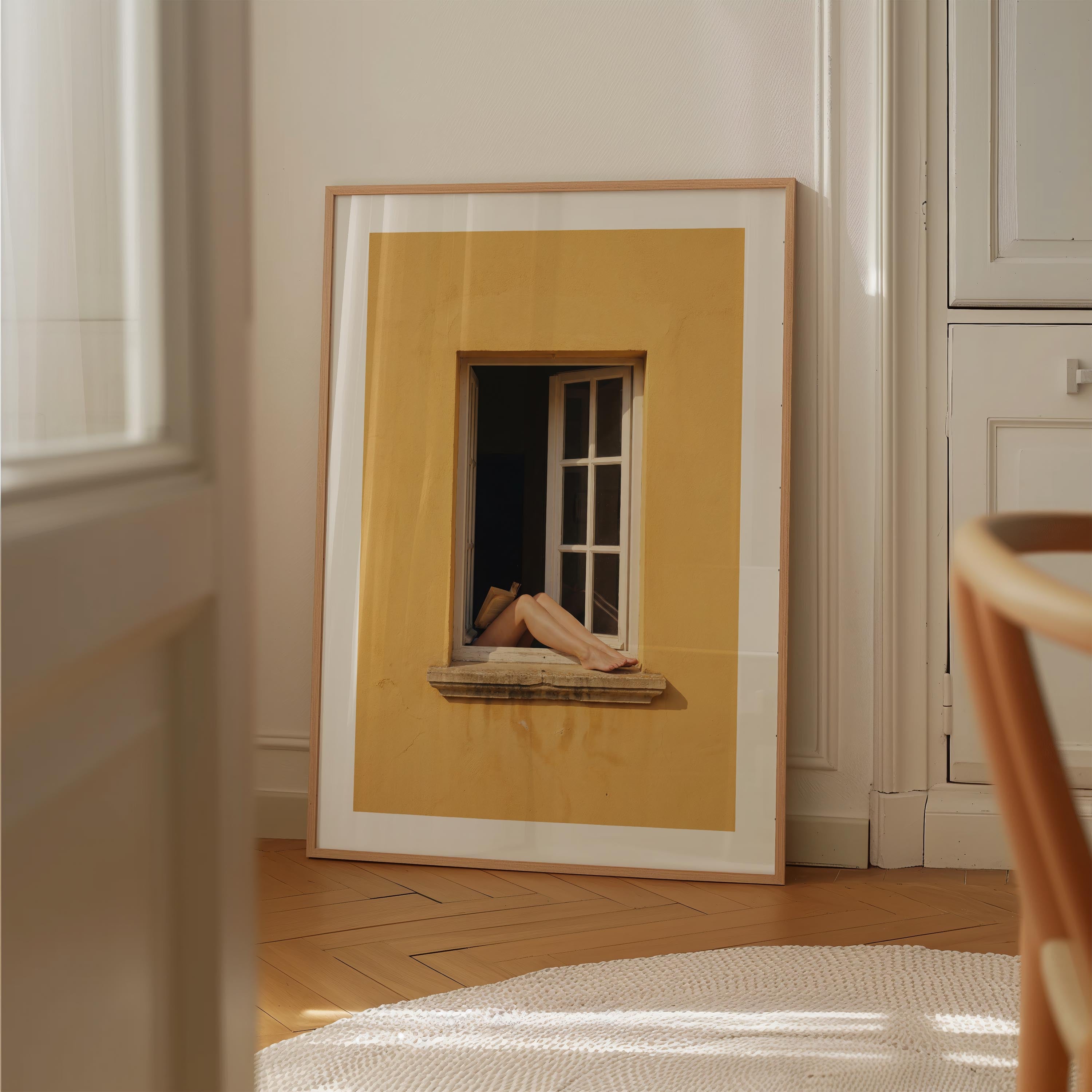 Art print showing bare legs and feet resting on the stone sill of an open white-framed window set into a warm golden-yellow Mediterranean wall, in a natural wood frame with white mat border.