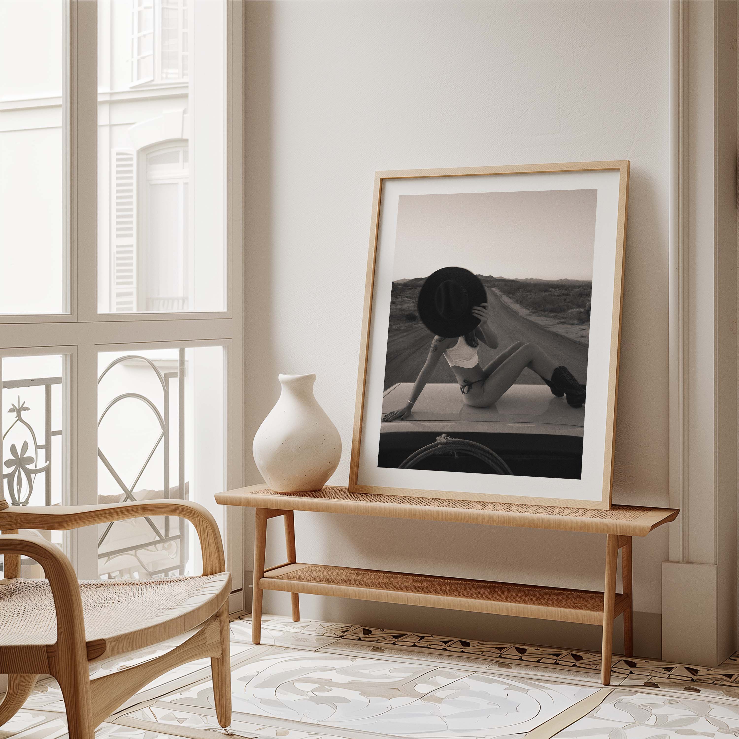 Fine art photo print of a woman sitting on a car roof on a desert road, holding a wide-brim hat in front of her face at sunset.