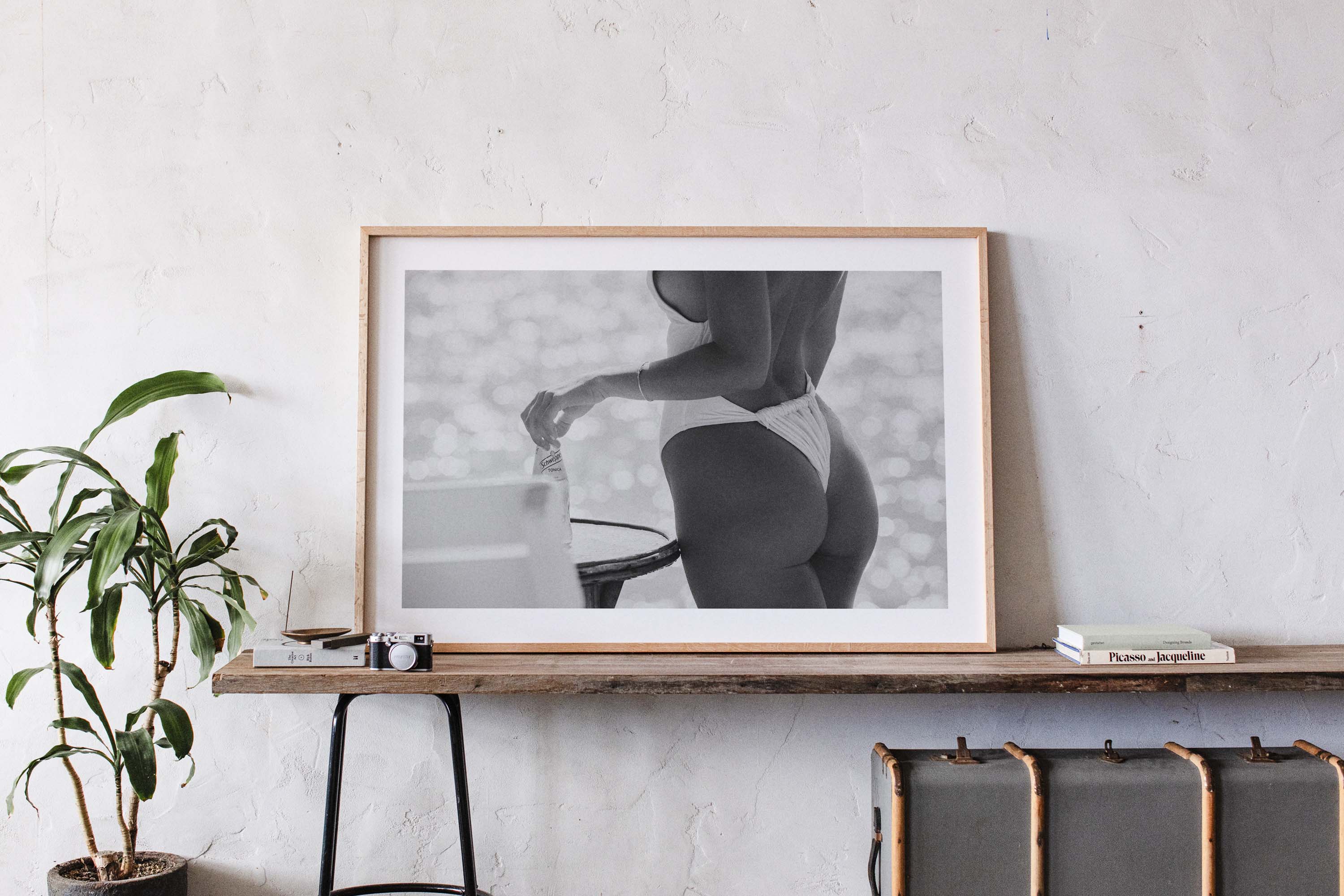 Black-and-white photo of a woman in a white one-piece holding a glass bottle by a sunlit seaside table