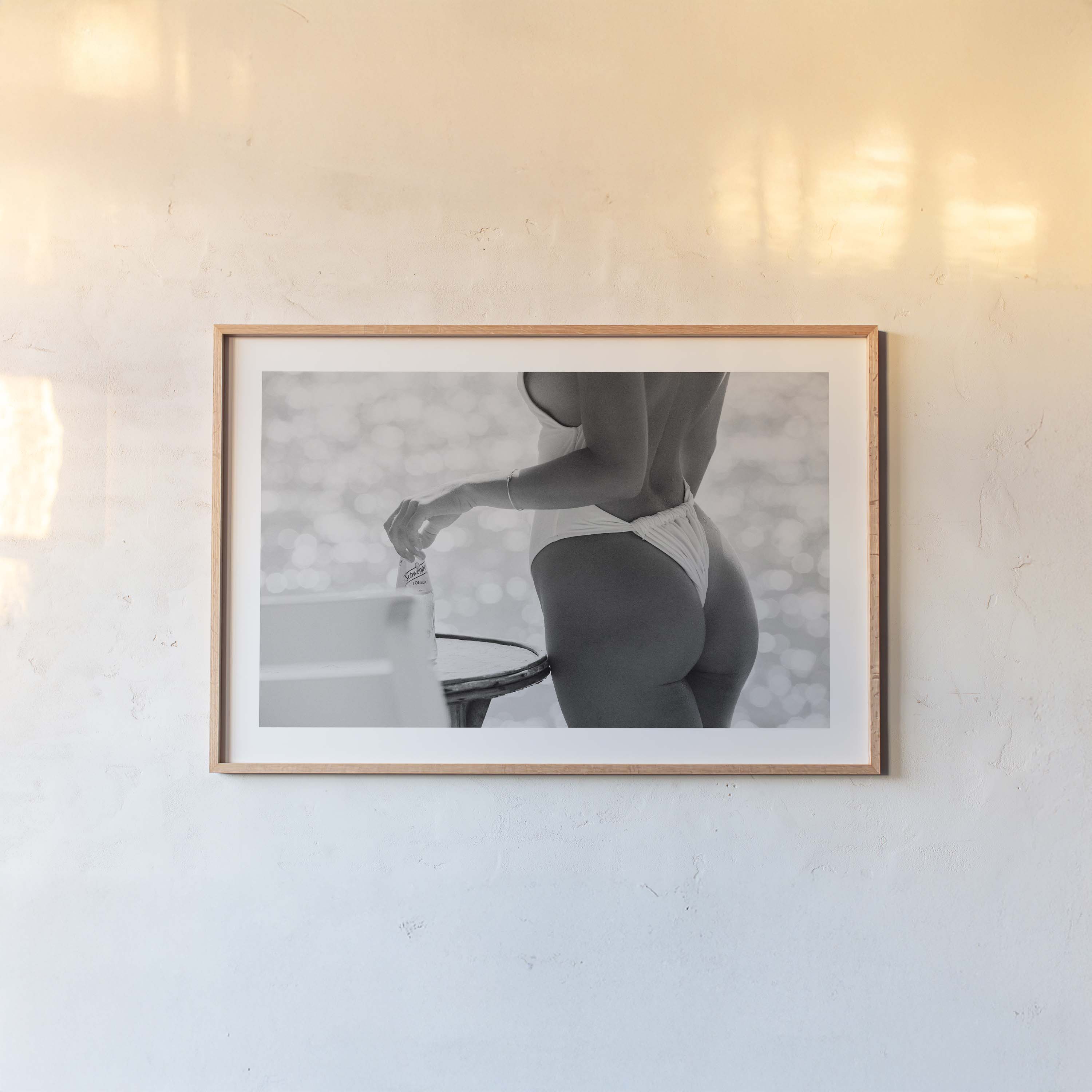 Black-and-white photo of a woman in a white one-piece holding a glass bottle by a sunlit seaside table