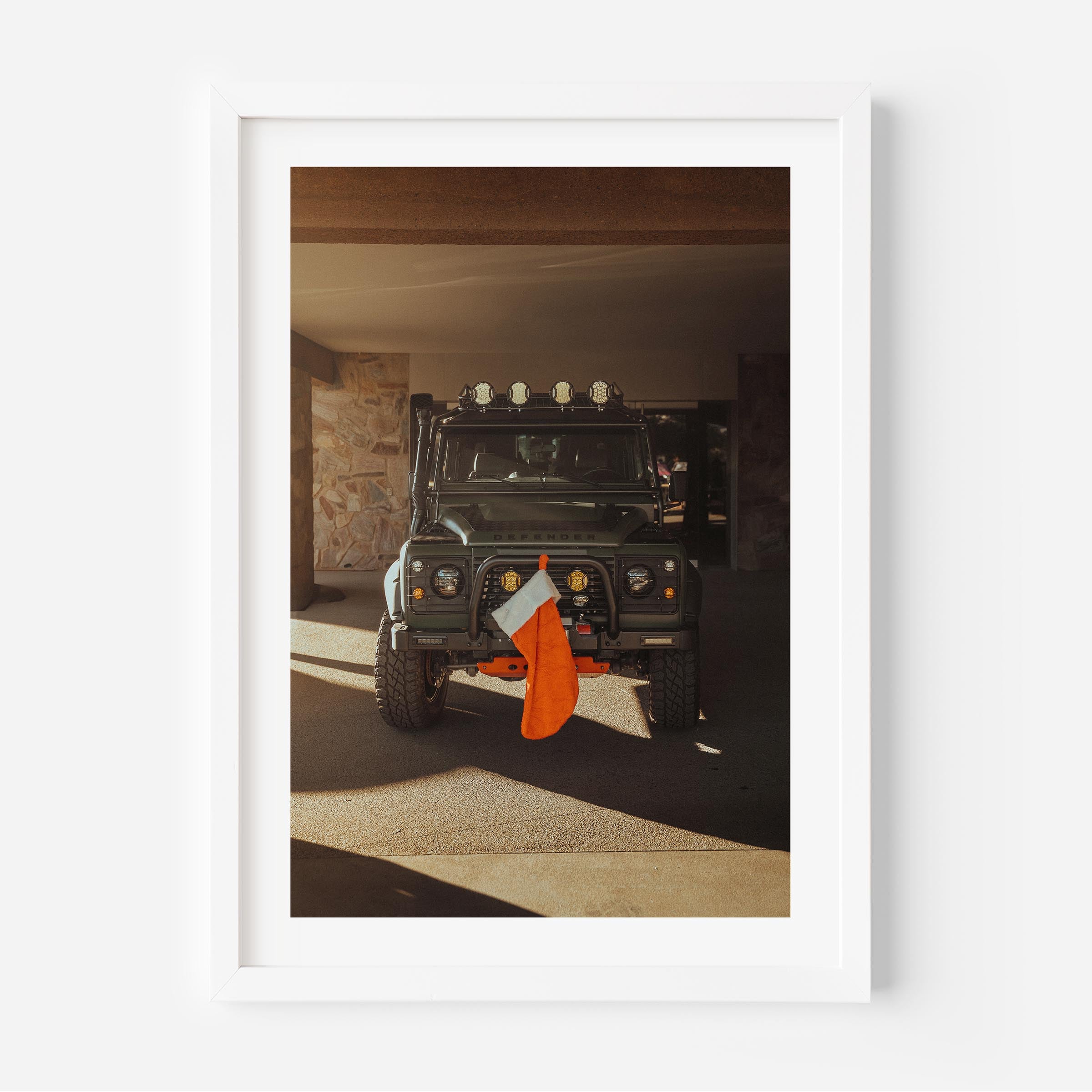 Fine art photo of a Land Rover Defender parked in a garage with warm sunlight, featuring a red Christmas stocking hanging from the front bumper.