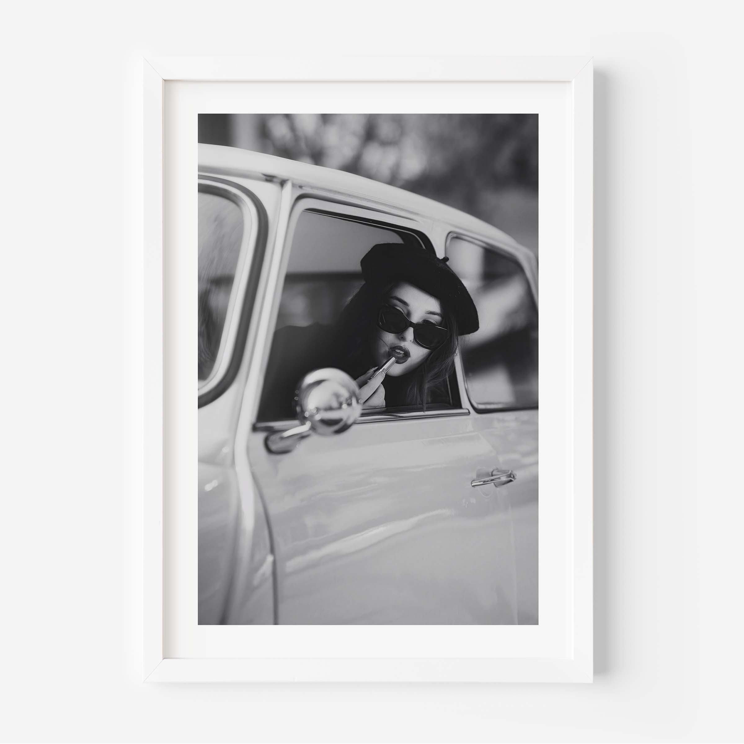 Black and white framed wall art print showing a woman in sunglasses sitting in a vintage car while applying lipstick.