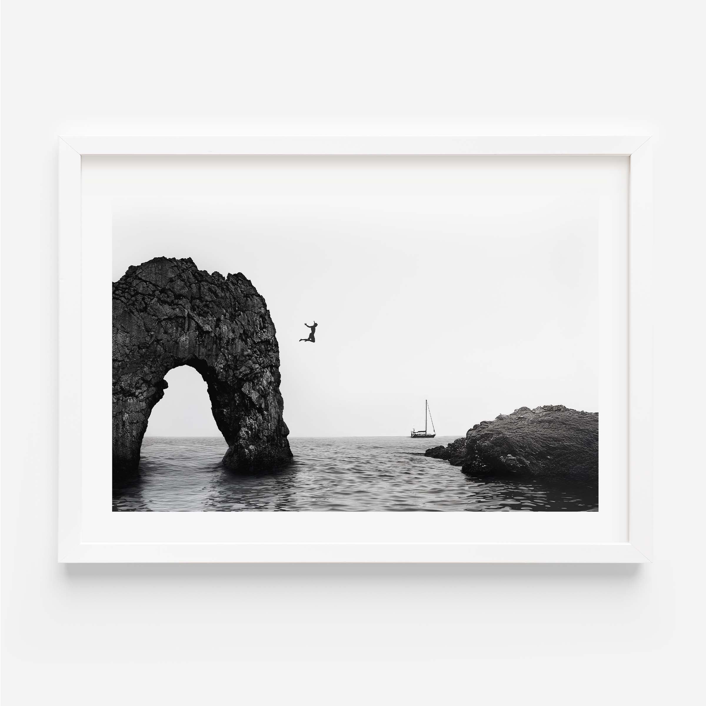 Black and white framed wall art print showing a person jumping into the ocean near a large rock arch with a sailboat in the distance.