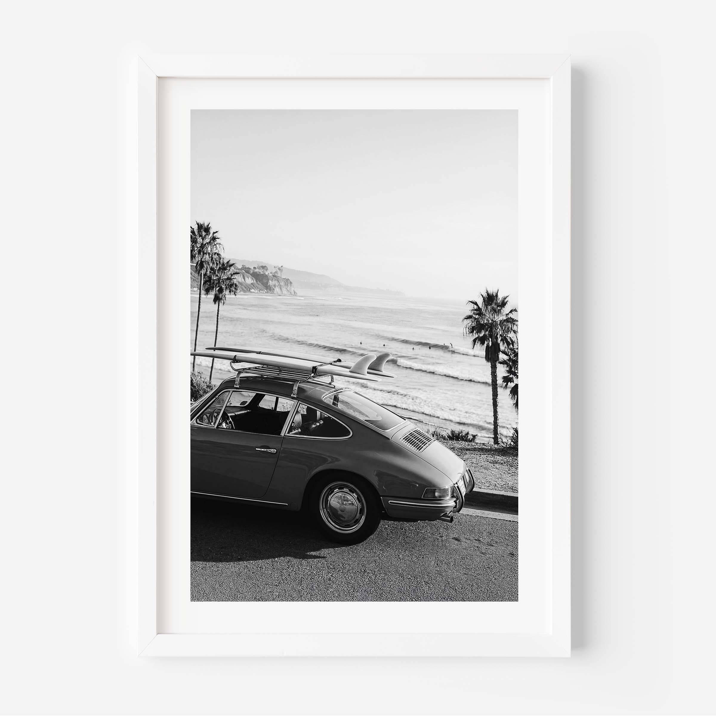 Black and white framed wall art print showing a classic porsche car with surfboards parked by the ocean with palm trees and coastline in the background.