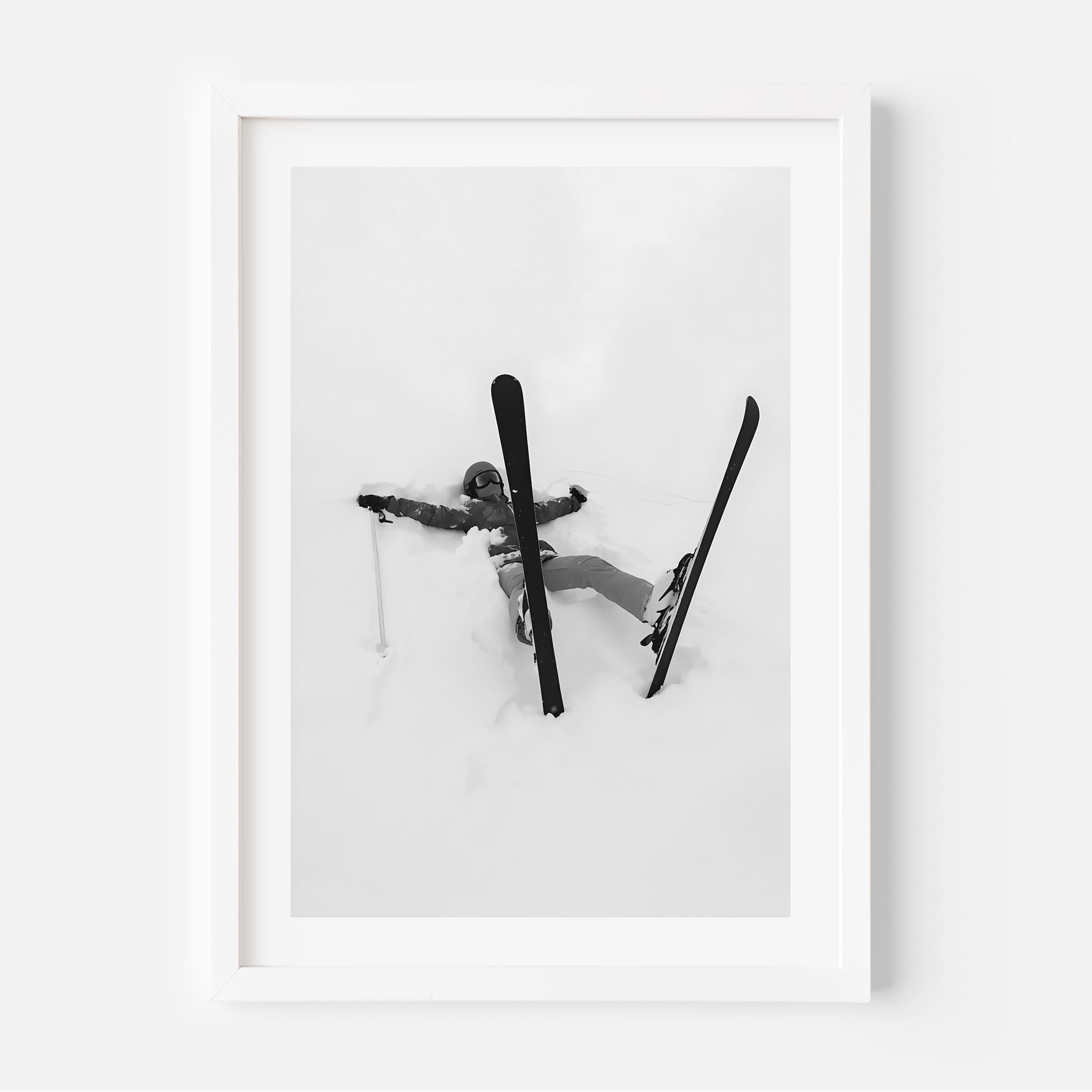 Black and white fine art print of a skier lying in deep snow with skis and pole scattered, framed in black.