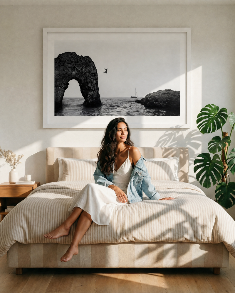 Woman sitting on a bed in a bright bedroom with a large framed photograph of a coastal scene above her.