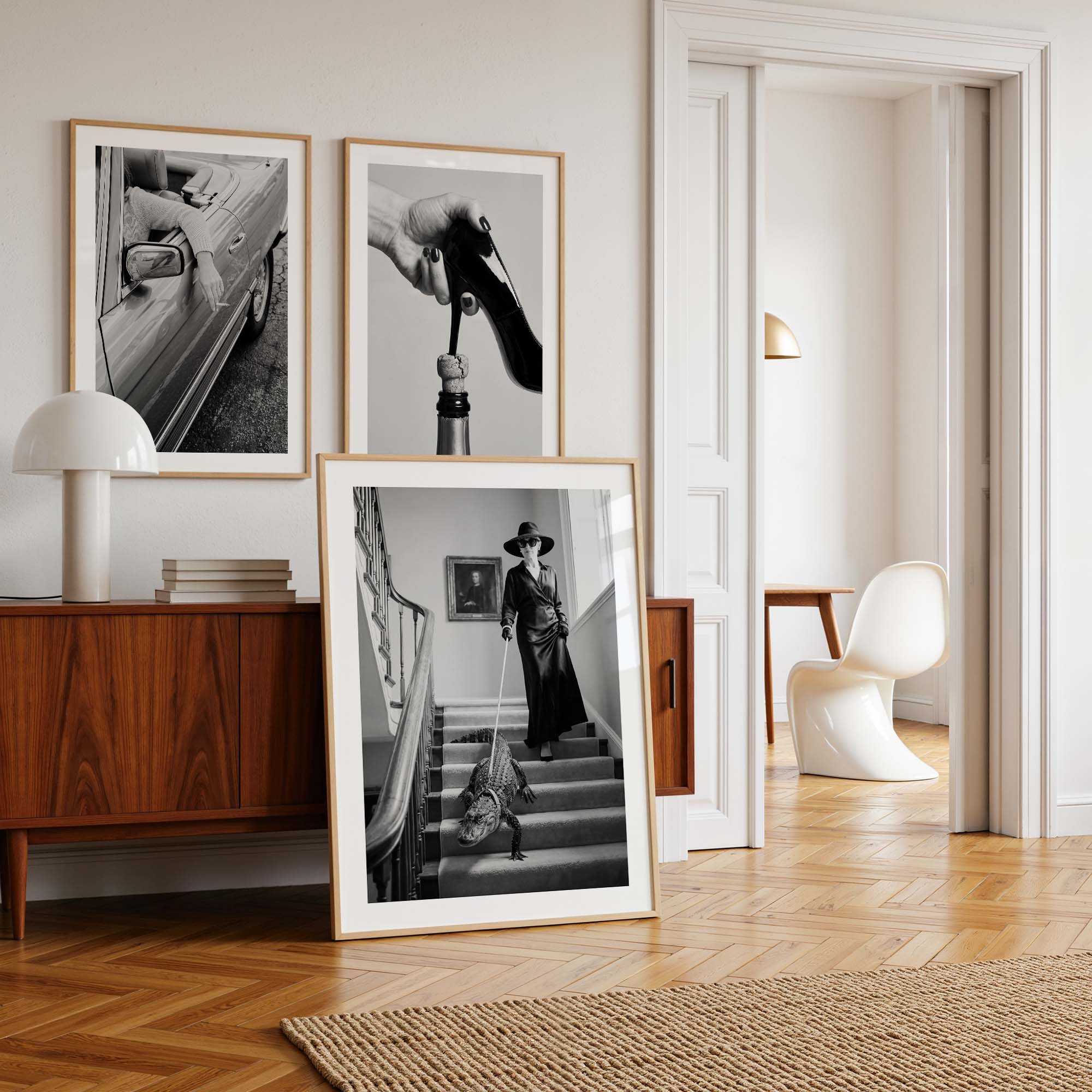 Three framed black and white art prints with light wood frames are displayed in a stylishly decorated room. Two prints are mounted on the wall above a mid-century modern wooden sideboard with a white table lamp, while the third print is propped up on the floor in front of the cabinet.