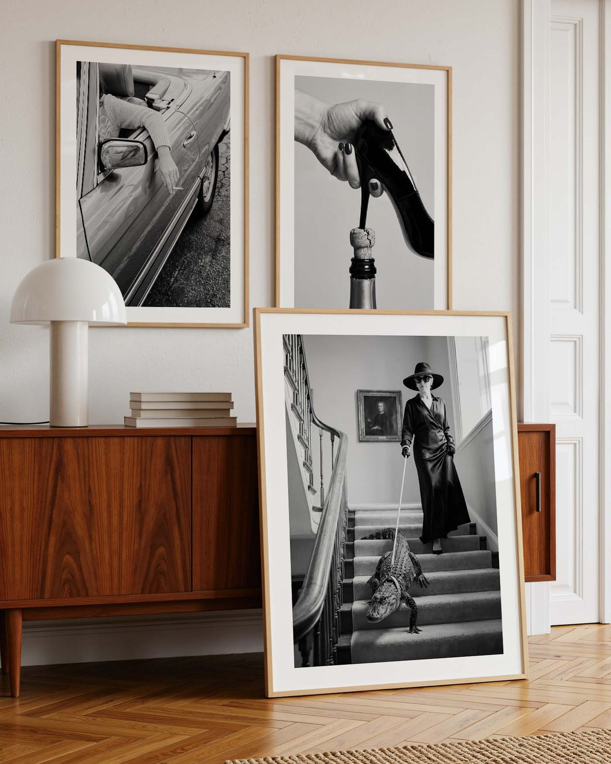 Three framed black and white art prints with light wood frames are displayed in a stylishly decorated room. Two prints are mounted on the wall above a mid-century modern wooden sideboard with a white table lamp, while the third print is propped up on the floor in front of the cabinet.