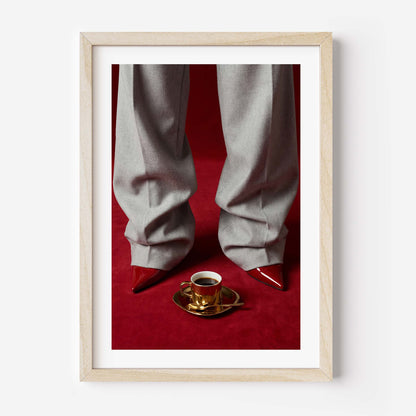 Fashion editorial wall art print showing grey wide-leg trousers, red pointed-toe stiletto heels and a gold espresso cup on a deep red velvet floor, in a black frame with white mat border.