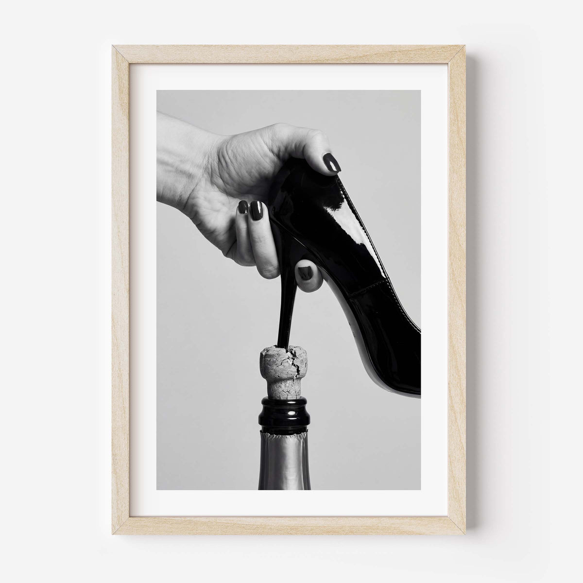Framed black and white art print featuring a hand holding a high-heeled shoe above a champagne bottle cork, in a light wood frame with a white mat.