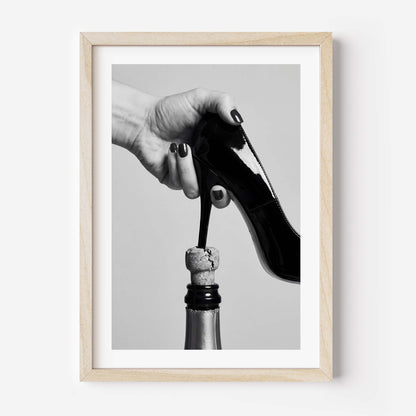 Framed black and white art print featuring a hand holding a high-heeled shoe above a champagne bottle cork, in a light wood frame with a white mat.