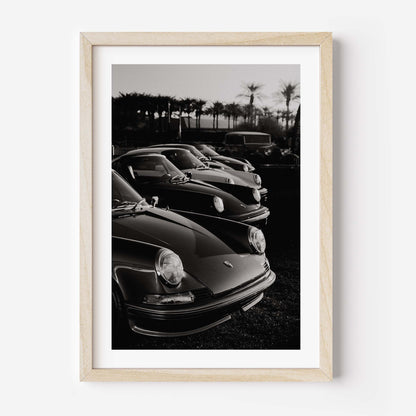 Black-and-white photo print of a row of classic Porsche 911 sports cars parked with palm trees in the background.