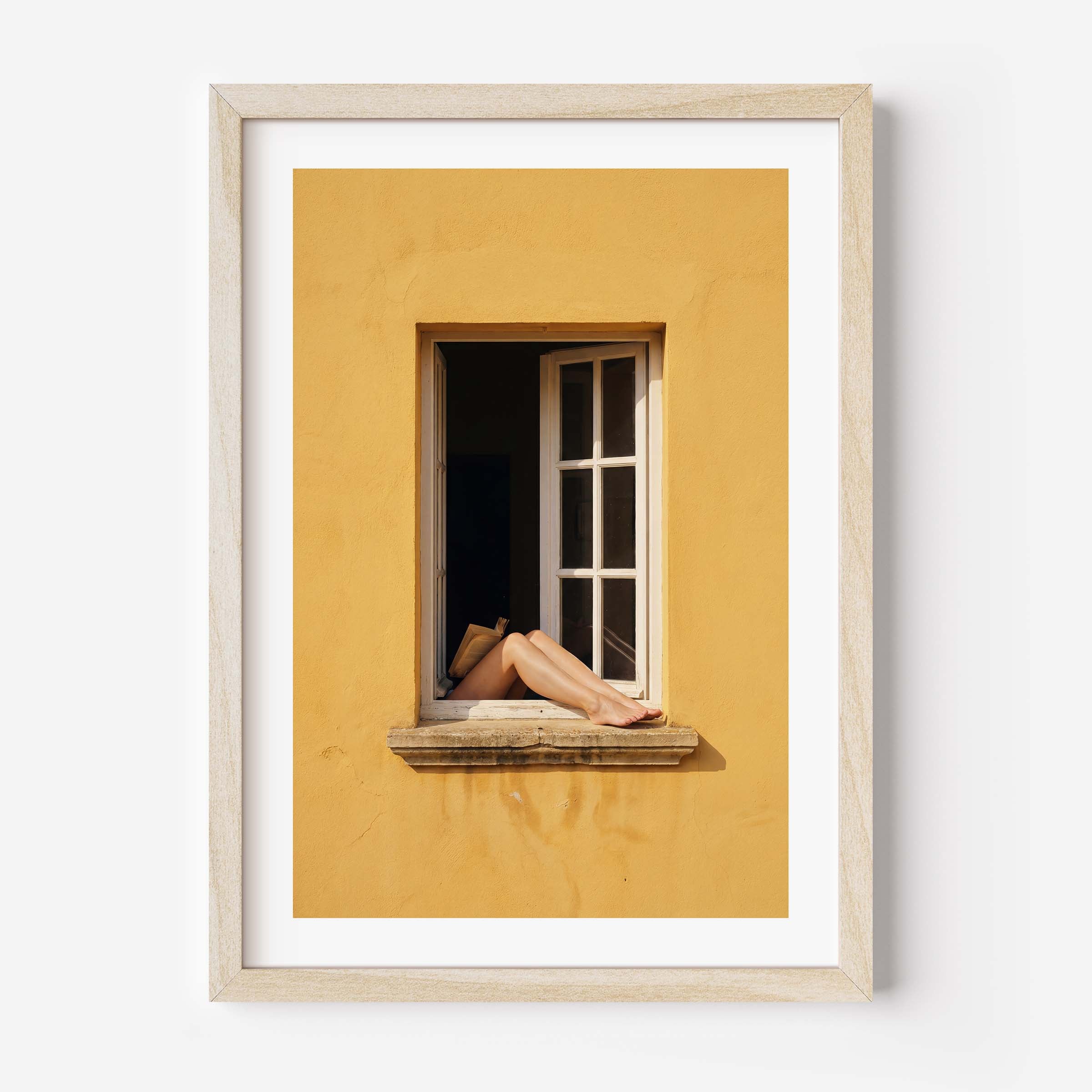 
Art print showing bare legs and feet resting on the stone sill of an open white-framed window set into a warm golden-yellow Mediterranean wall, in a natural wood frame with white mat border.