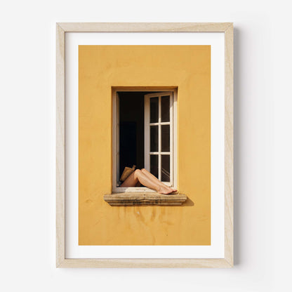 
Art print showing bare legs and feet resting on the stone sill of an open white-framed window set into a warm golden-yellow Mediterranean wall, in a natural wood frame with white mat border.