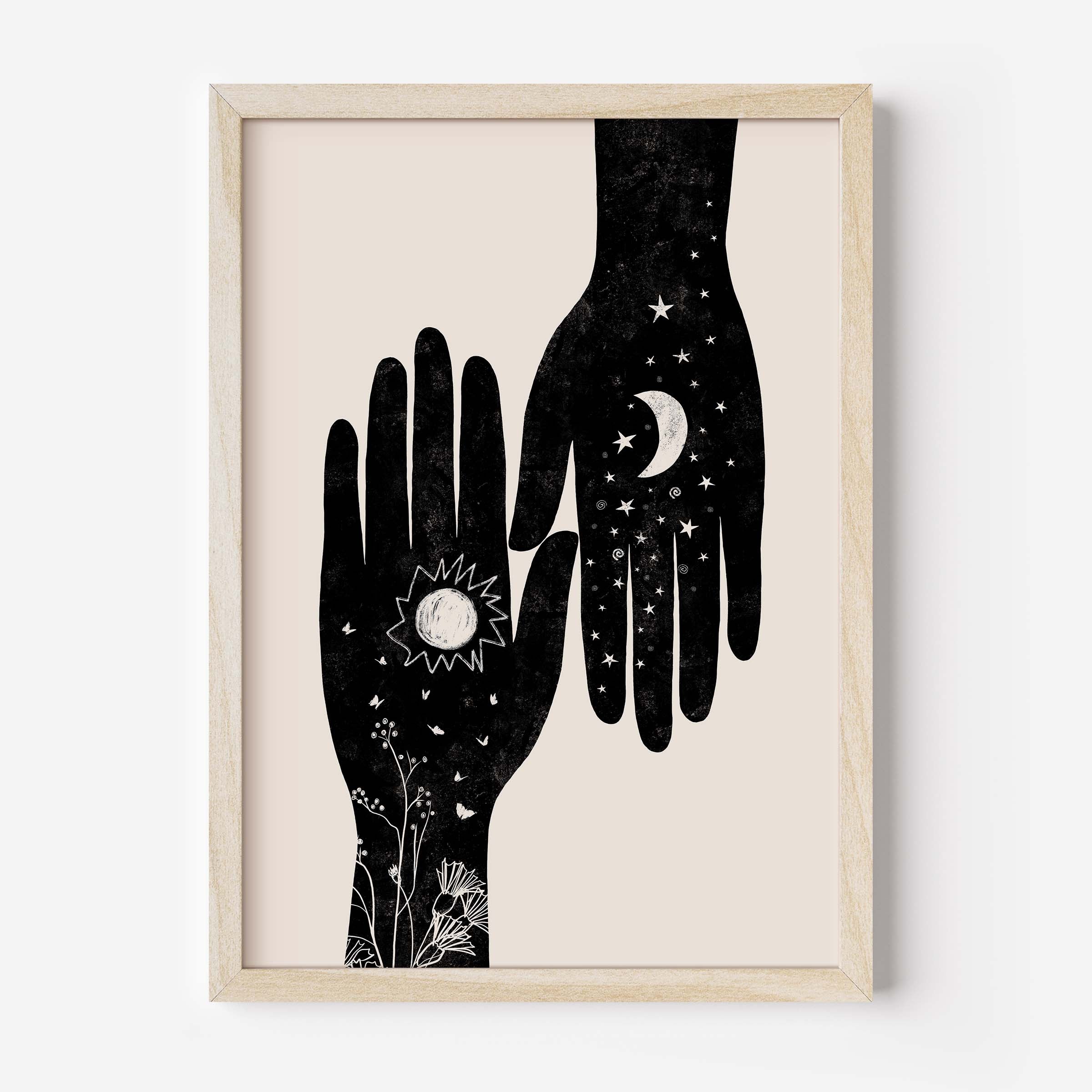 Framed wall art featuring a minimalist black illustration of two hands with celestial and floral details on a warm cream background, set in a light natural wood frame.
