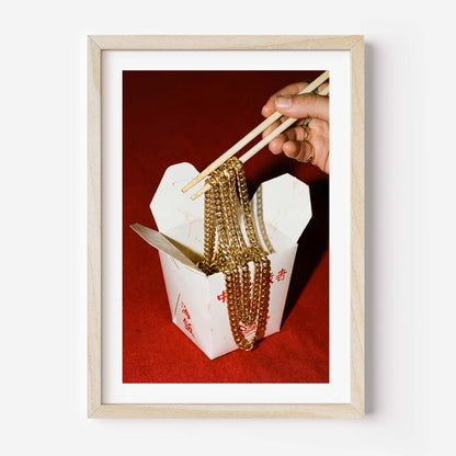 Conceptual photography print of a hand lifting gold chains with chopsticks from a Chinese takeout box on a red background, framed in black